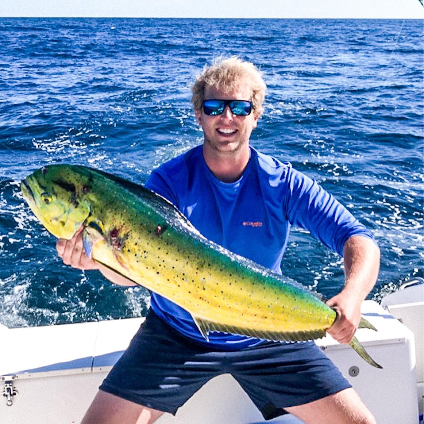 Man in sunglasses holds a large, colorful mahi-mahi fish on a boat, ocean in the background.