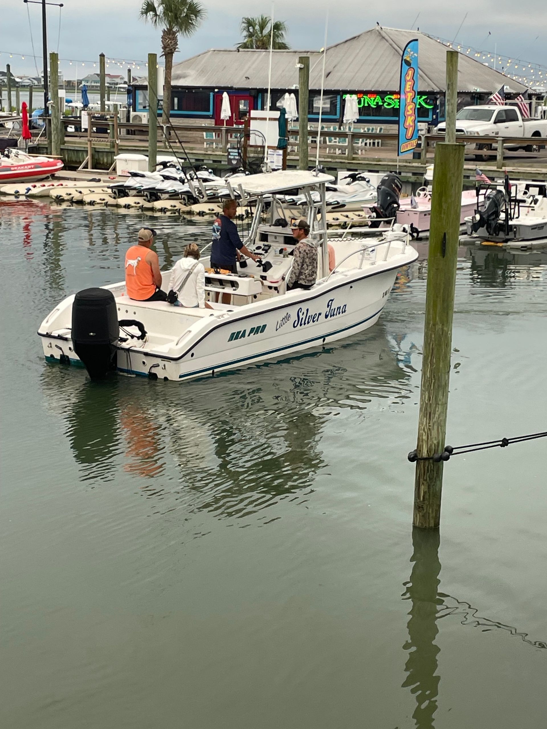 Boat with three people in harbor. White boat with blue text, near dock and buildings.