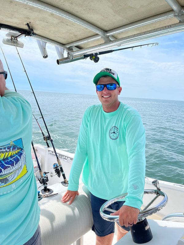 Man smiling at the helm of a boat, wearing sunglasses and a green shirt, with fishing rods in the background.