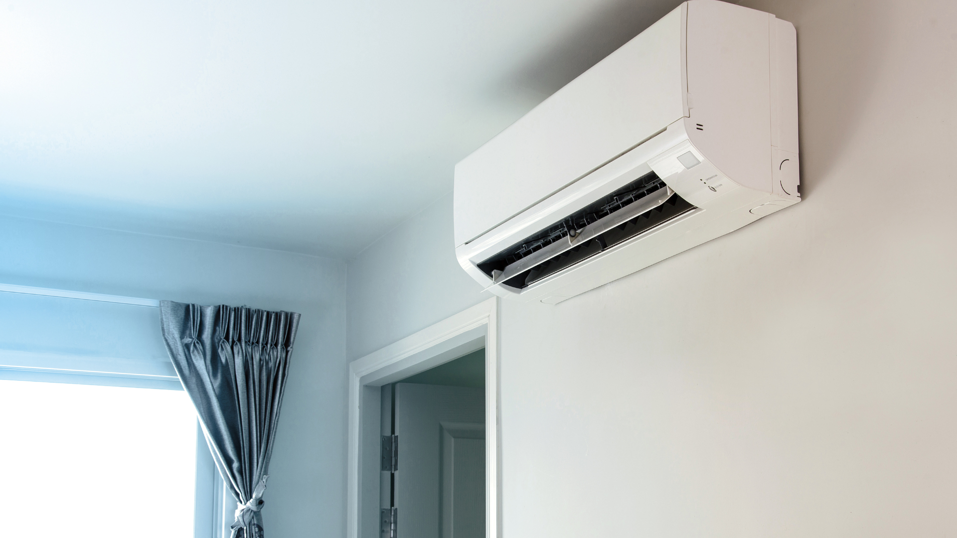 A wall-mounted white air conditioner unit in a room with a light wall and a window with striped curtains.