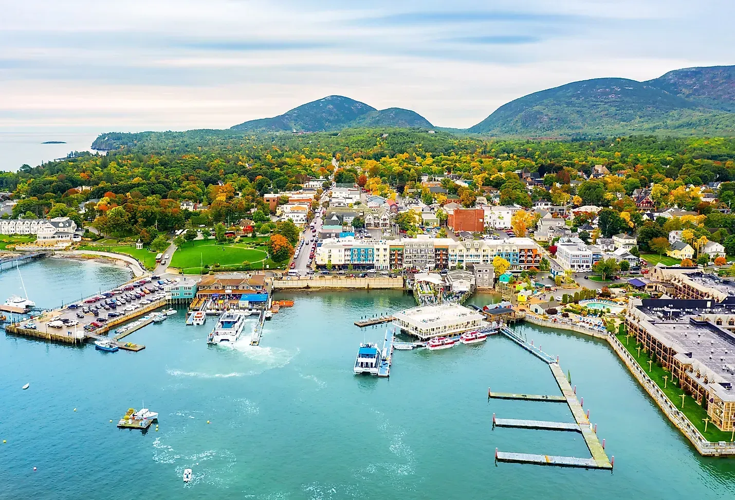Aerial view of harbor town: boats, docks, buildings, lush trees, and hills in the background under a cloudy sky.