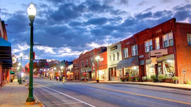 Street lined with brick buildings, illuminated storefronts, and streetlights under a twilight sky.