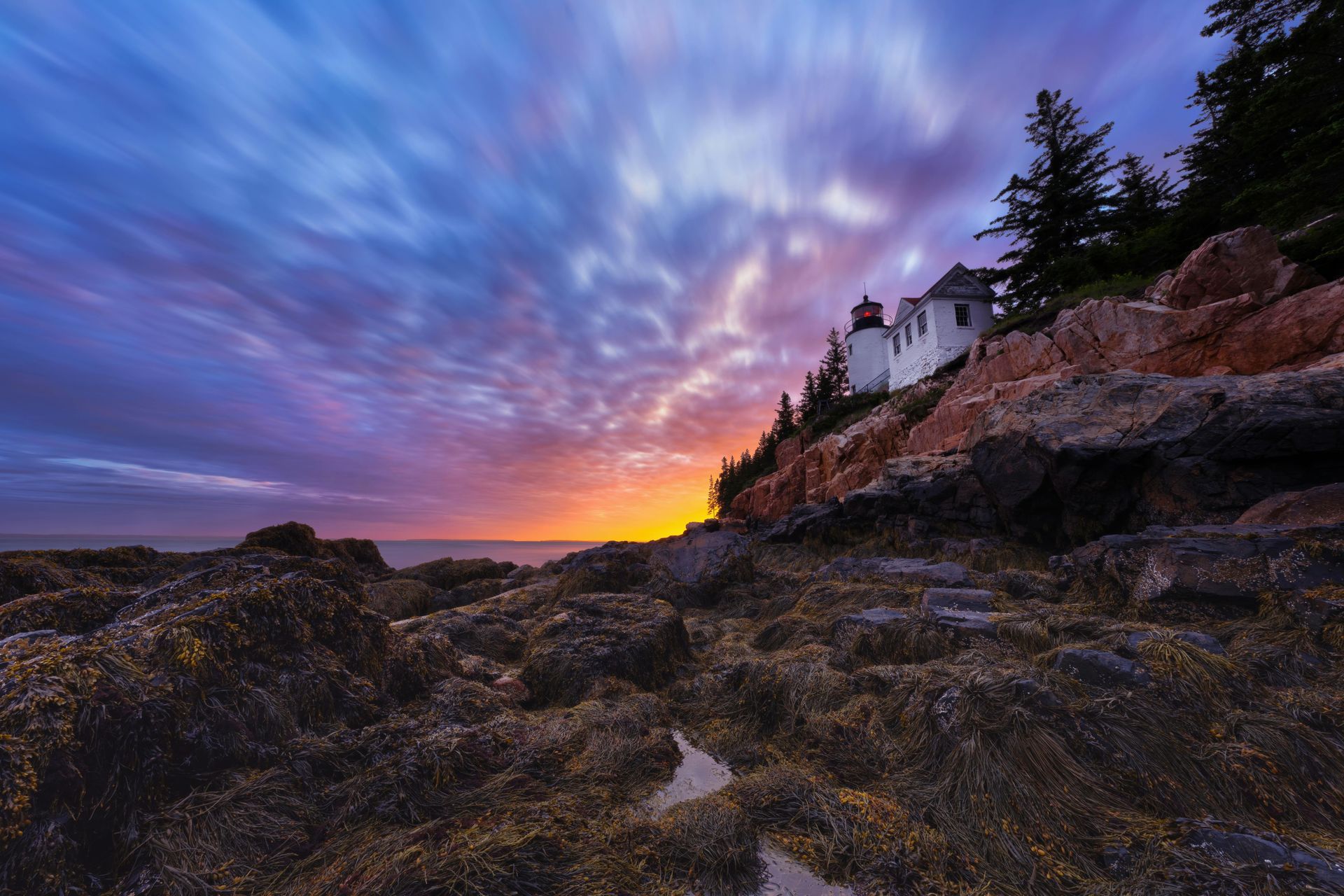 Lighthouse on a rocky coast at sunrise, with streaked blue and orange sky overhead.