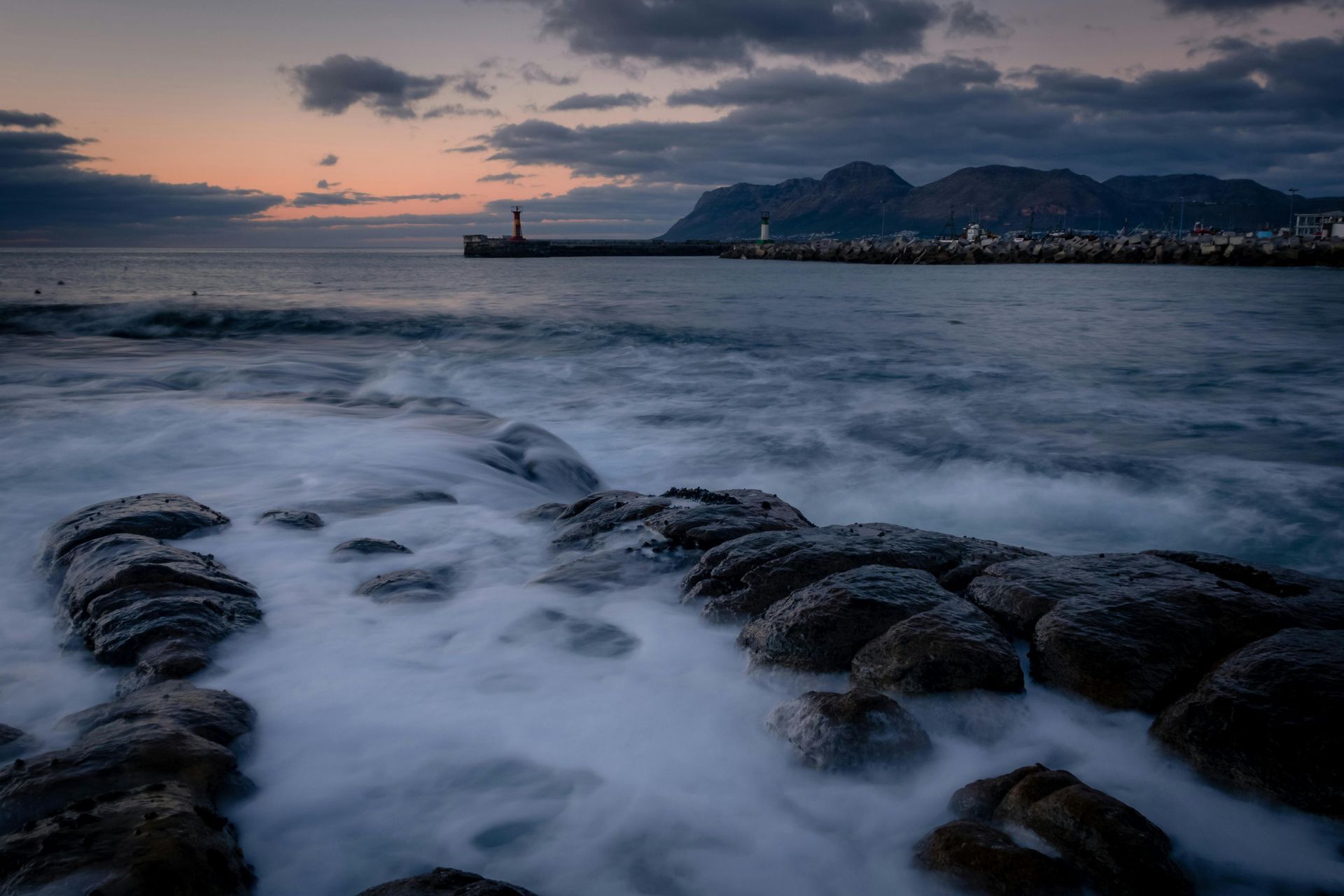 Rocky coastline with ocean waves at dusk, a harbor with lights in the distance.