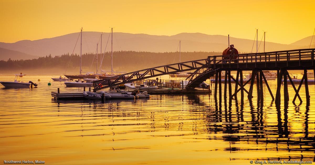 Rocky coastline with ocean waves at dusk, a harbor with lights in the distance.