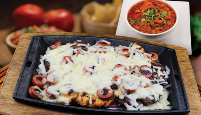 Sizzling platter with melted cheese, sausage, and meat; small bowl of stew on a wooden surface.
