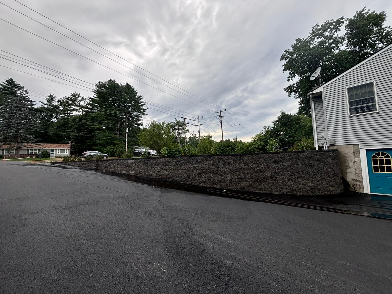 Asphalt road next to a dark brick retaining wall; house and cloudy sky in background.
