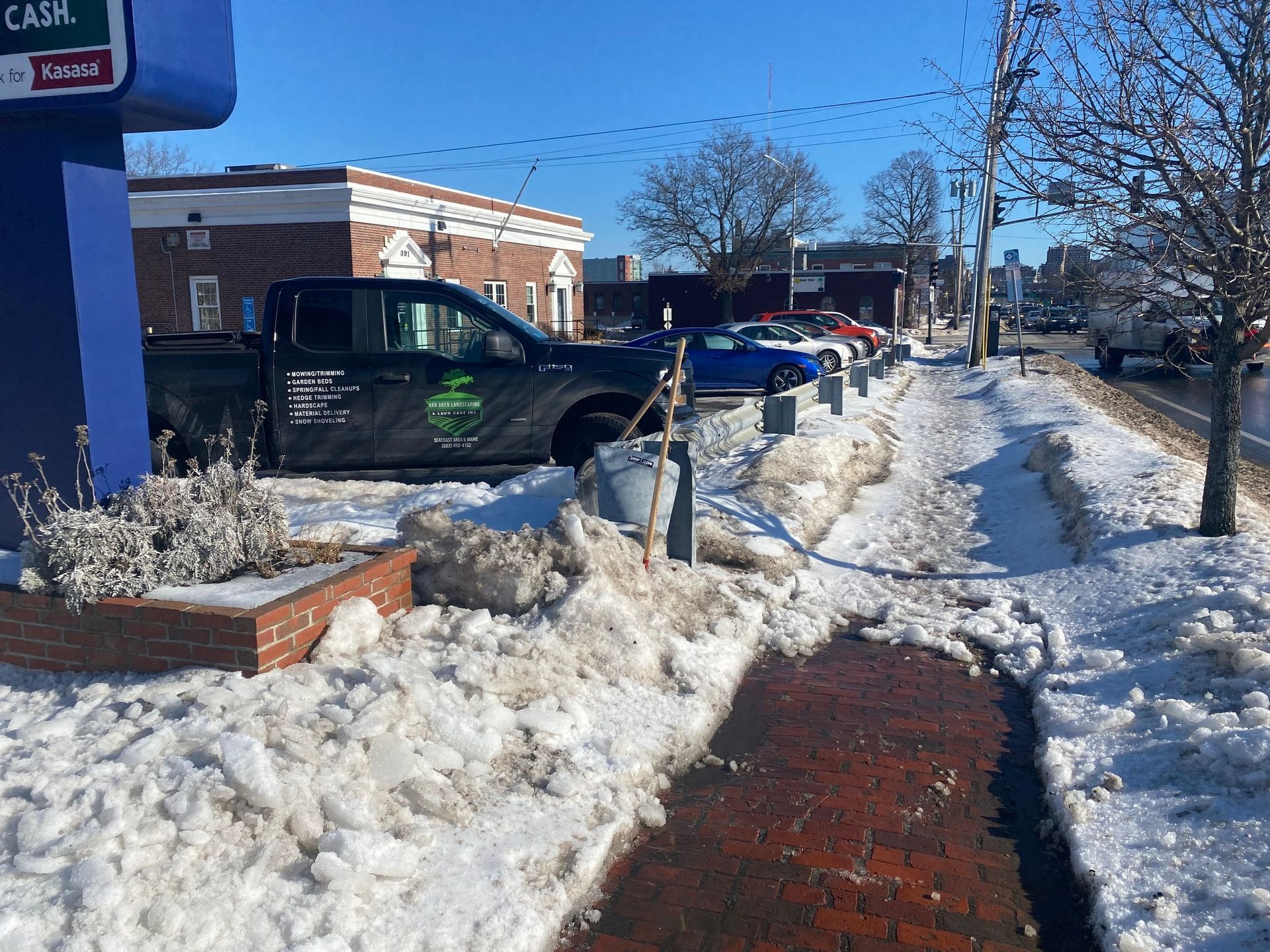 Sidewalk with packed snow and slush next to a street. Dark truck parked near building, a bucket on the sidewalk.