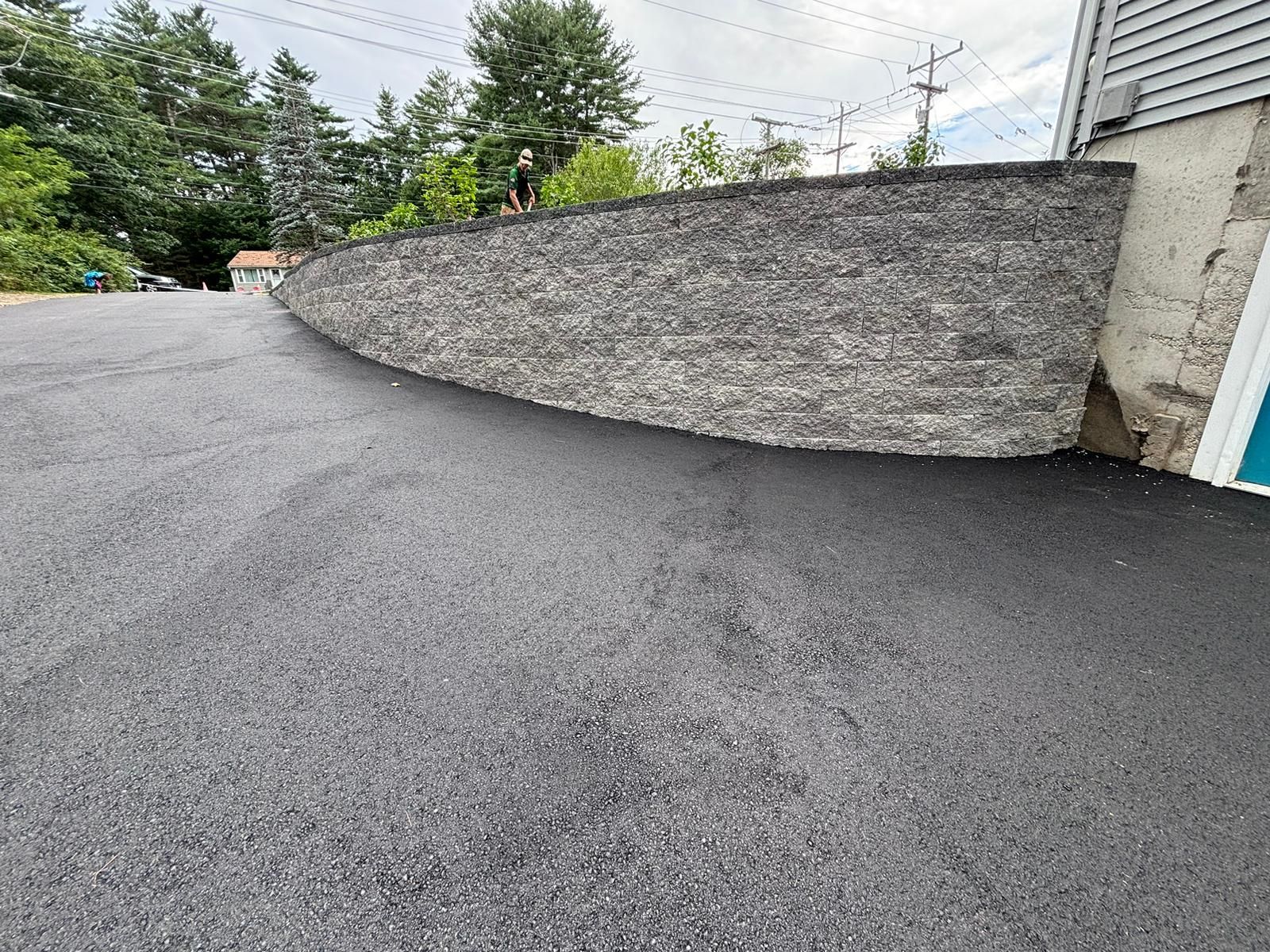 Asphalt driveway curving alongside a gray retaining wall next to a building. Trees in the background.