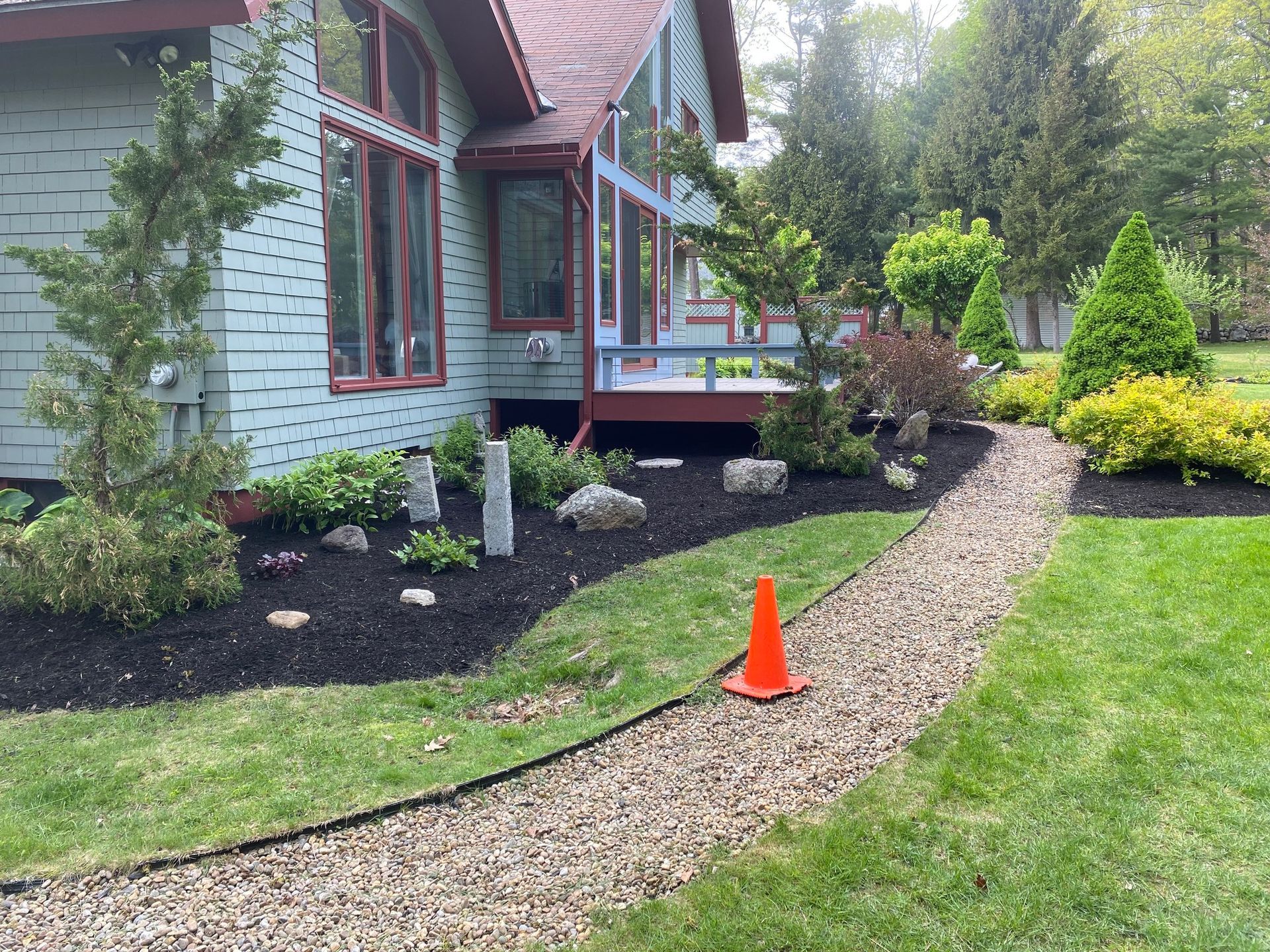 A gravel path curves through a landscaped yard with a house, mulch, plants, and a traffic cone.