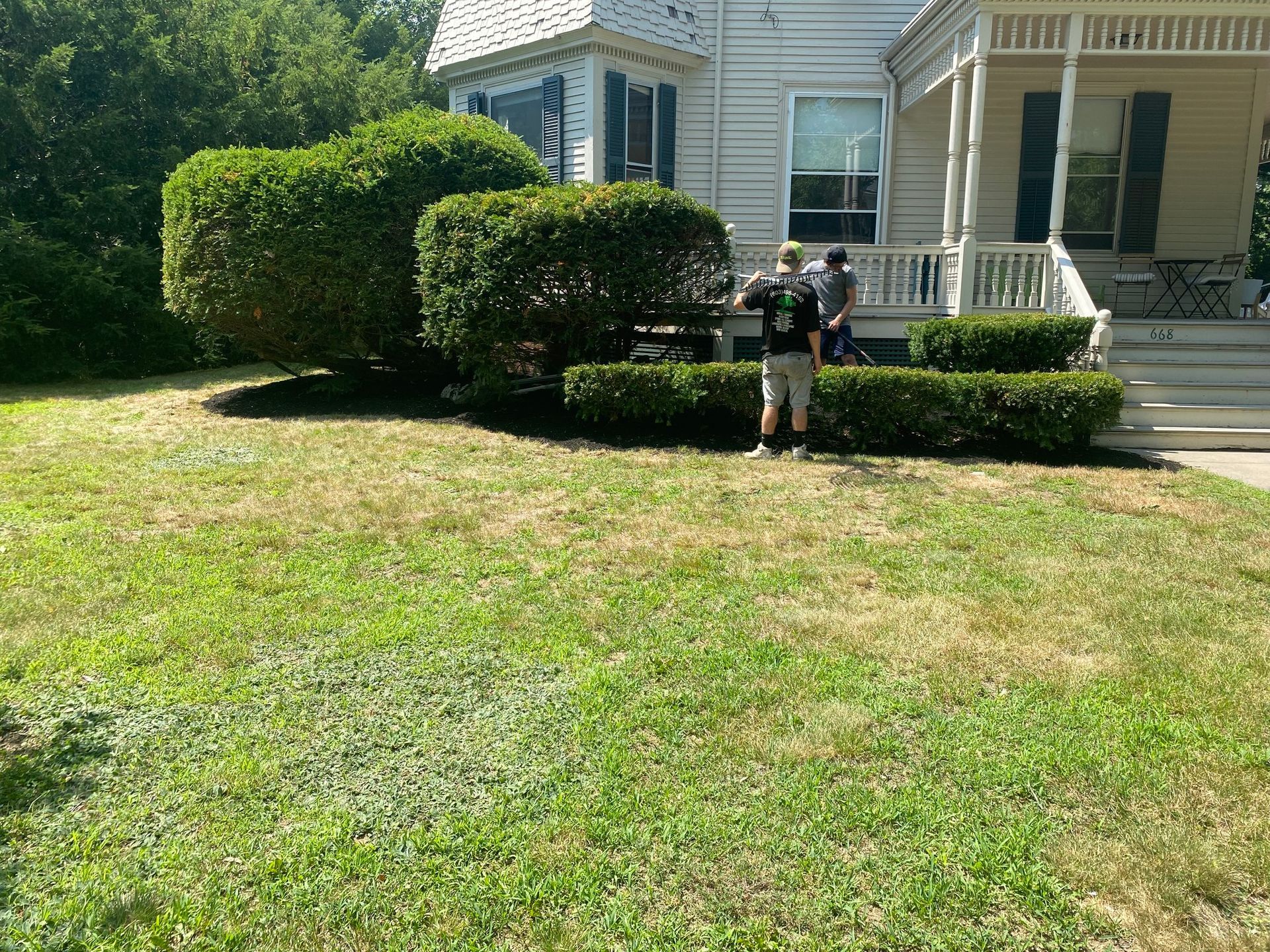 Two people trimming hedges in front of a white house with a porch and green lawn on a sunny day.