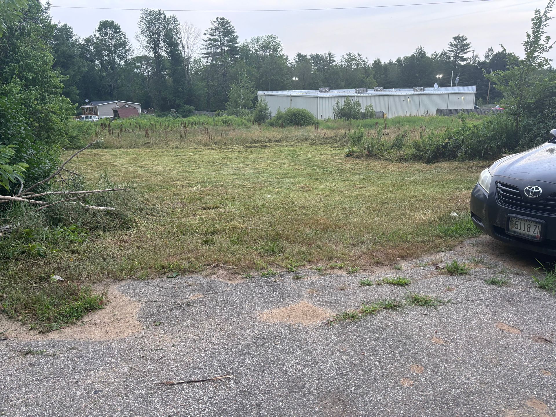 Gravel parking area in front of an overgrown grassy lot, car parked on right, buildings and trees in the background.
