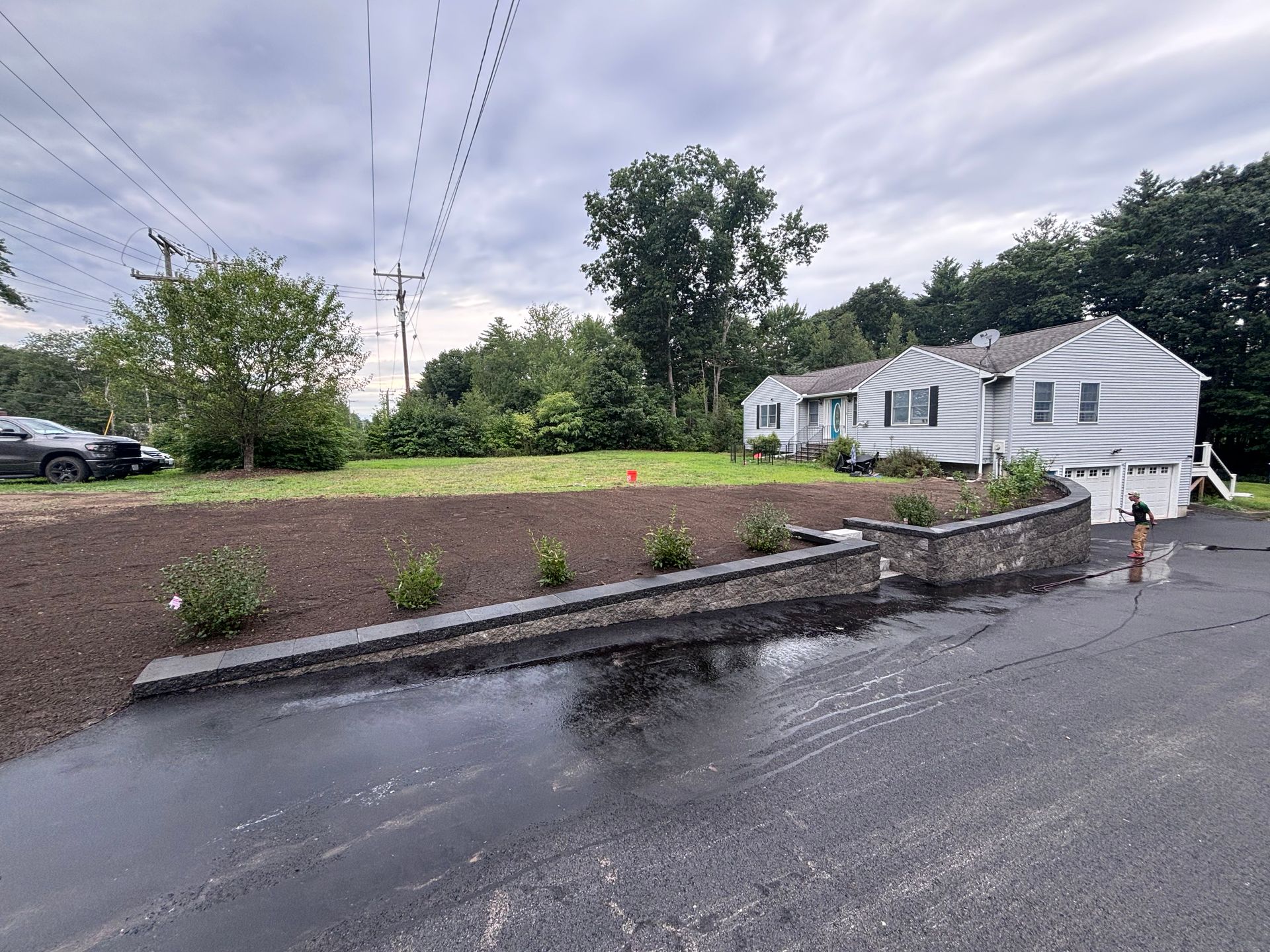 House with a gray stone retaining wall, newly planted shrubs, and a freshly paved driveway on an overcast day.