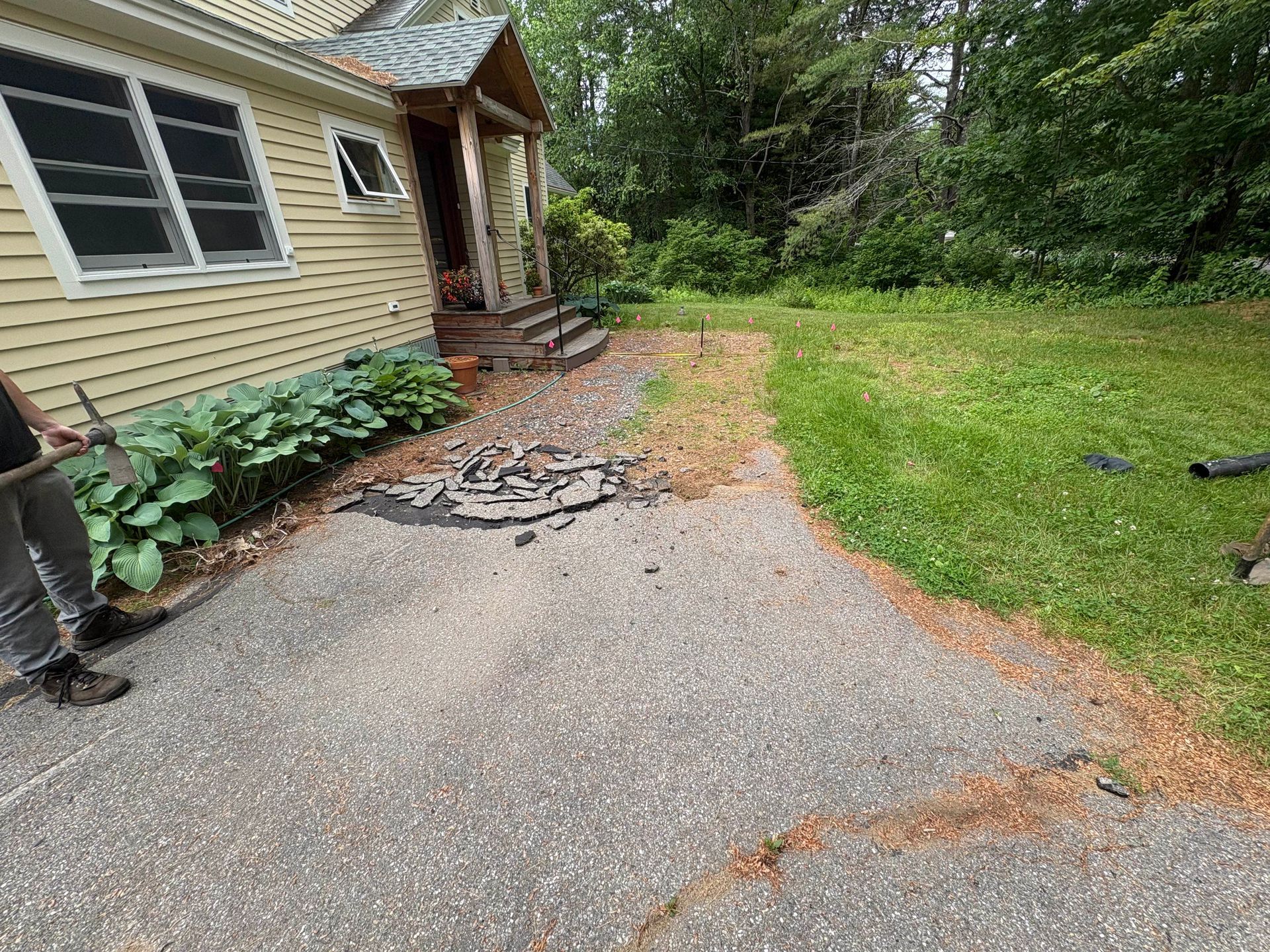 Driveway leading to a yellow house with a porch and steps, flanked by a lawn and overgrown landscaping.