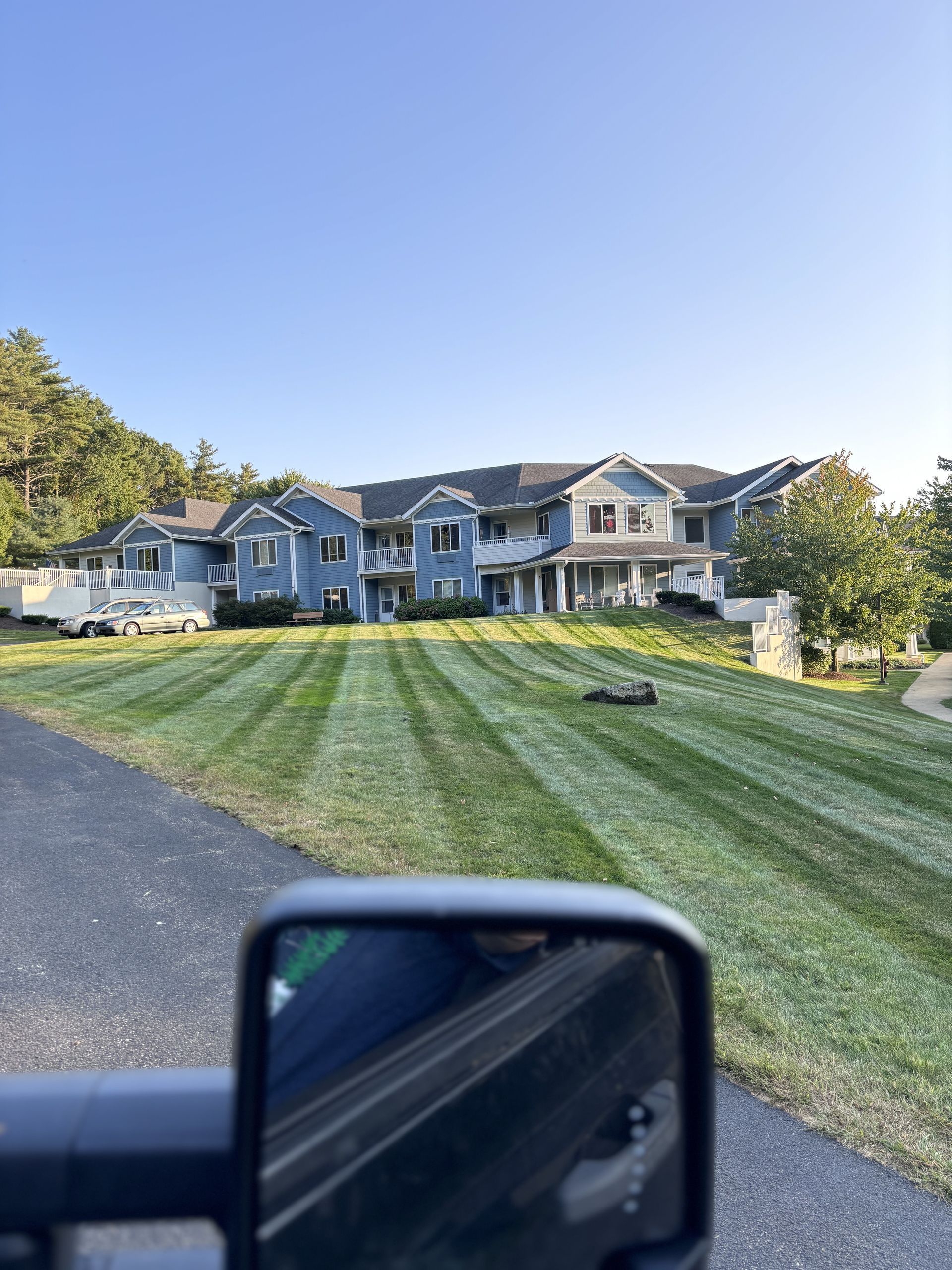 Large blue house with striped green lawn viewed through a vehicle's side mirror on a sunny day.