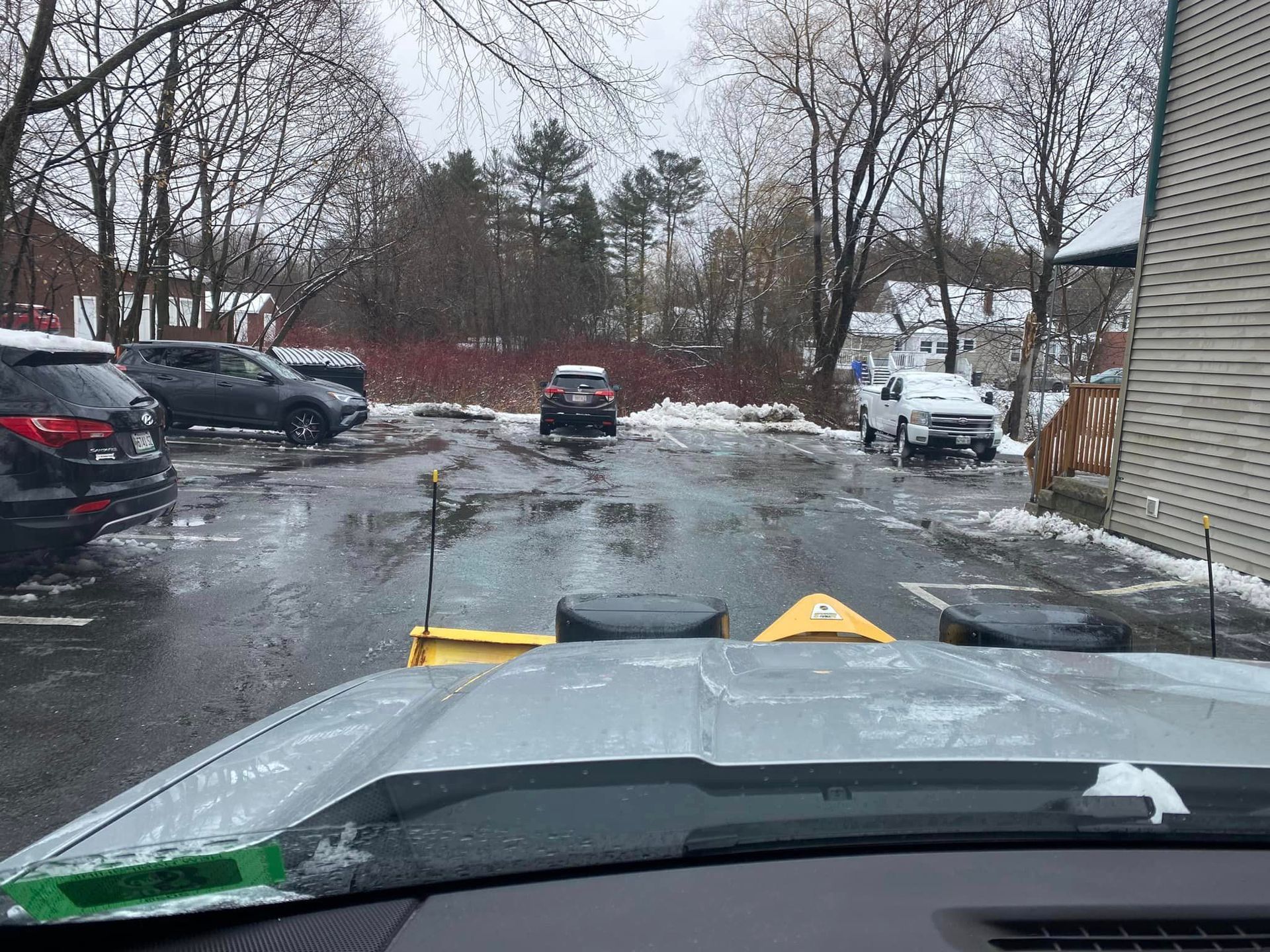 Snow-covered driveway leading to a house. Partially cleared of snow, chain link fence, and trees in the background.