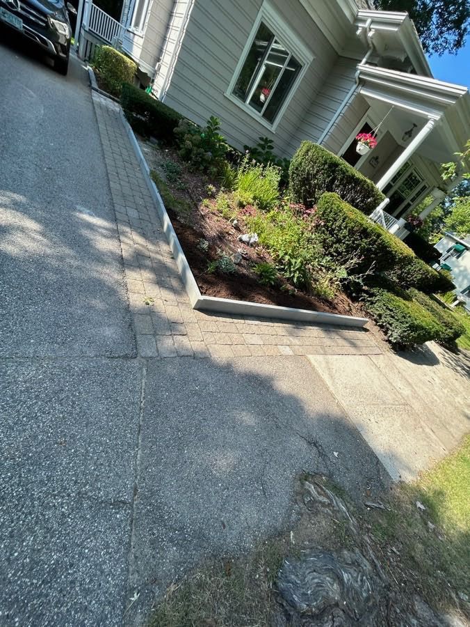 Asphalt driveway leading to a house with shrubs and flowers bordering the walkway.