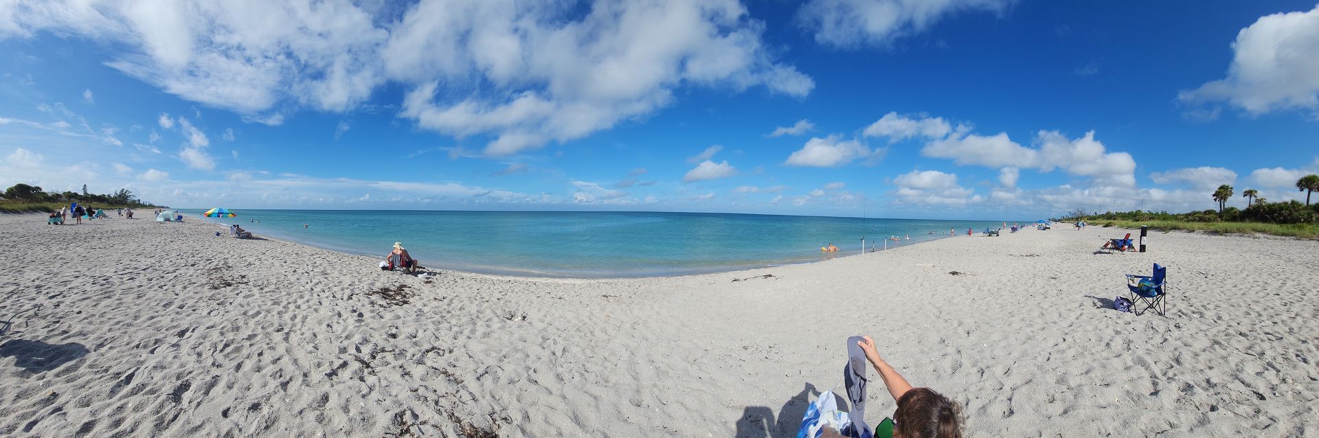 Wide sandy beach with scattered people, blue ocean, and a bright sky with clouds