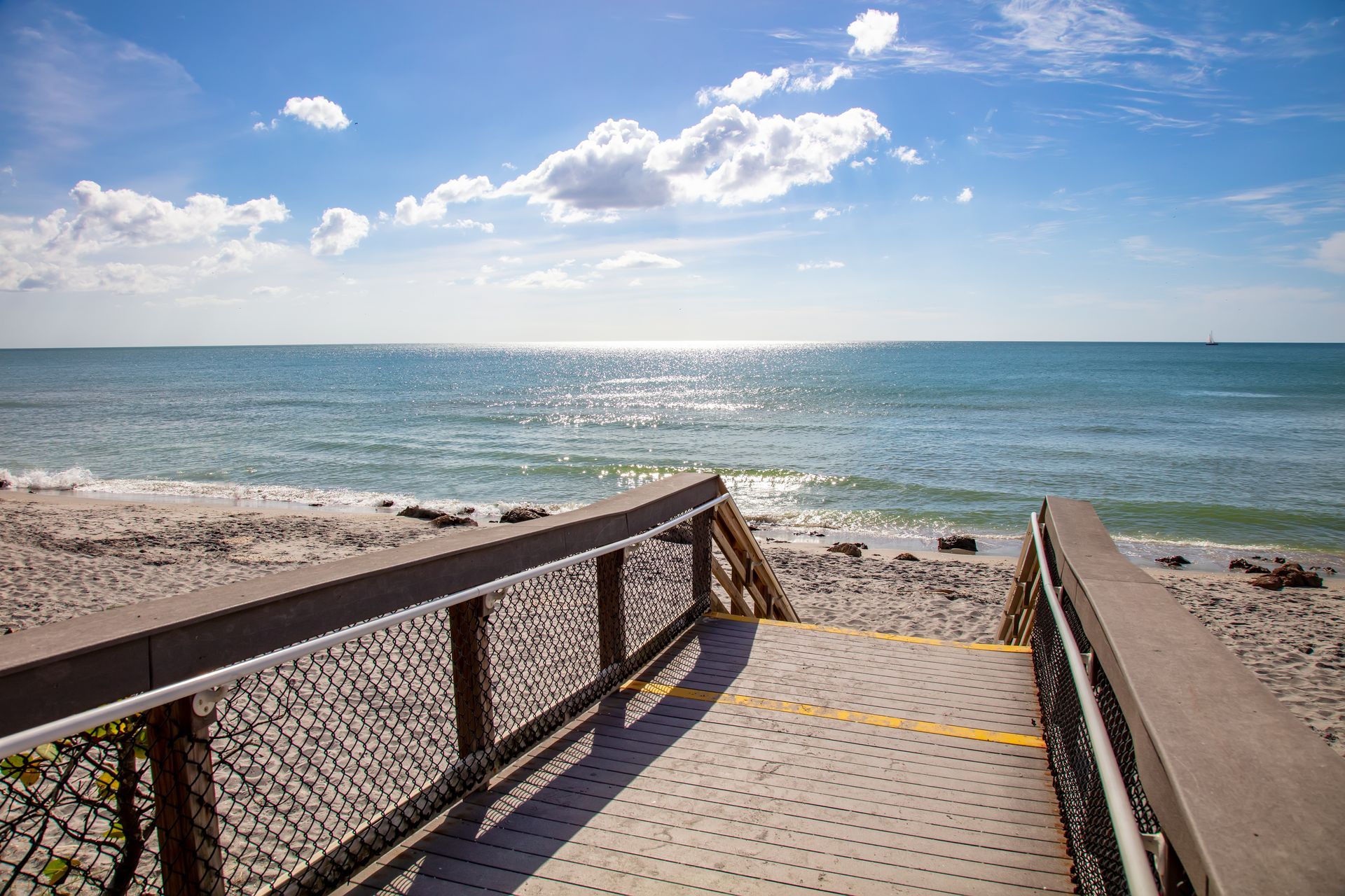Wooden beach walkway leading to a sunny ocean shoreline under a blue sky with clouds