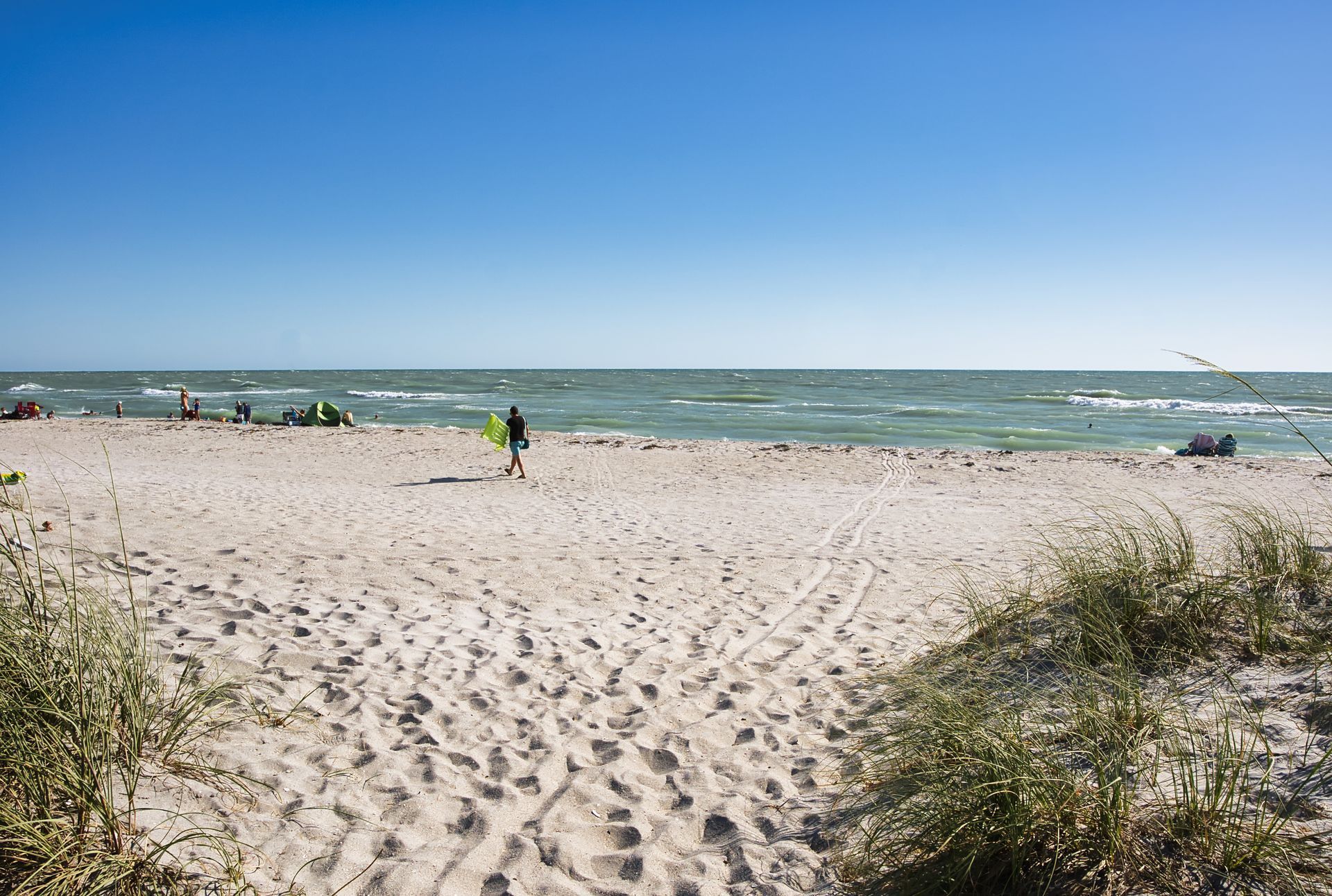 Sandy beach with dunes, a person near the shoreline, and a blue sky over choppy water.