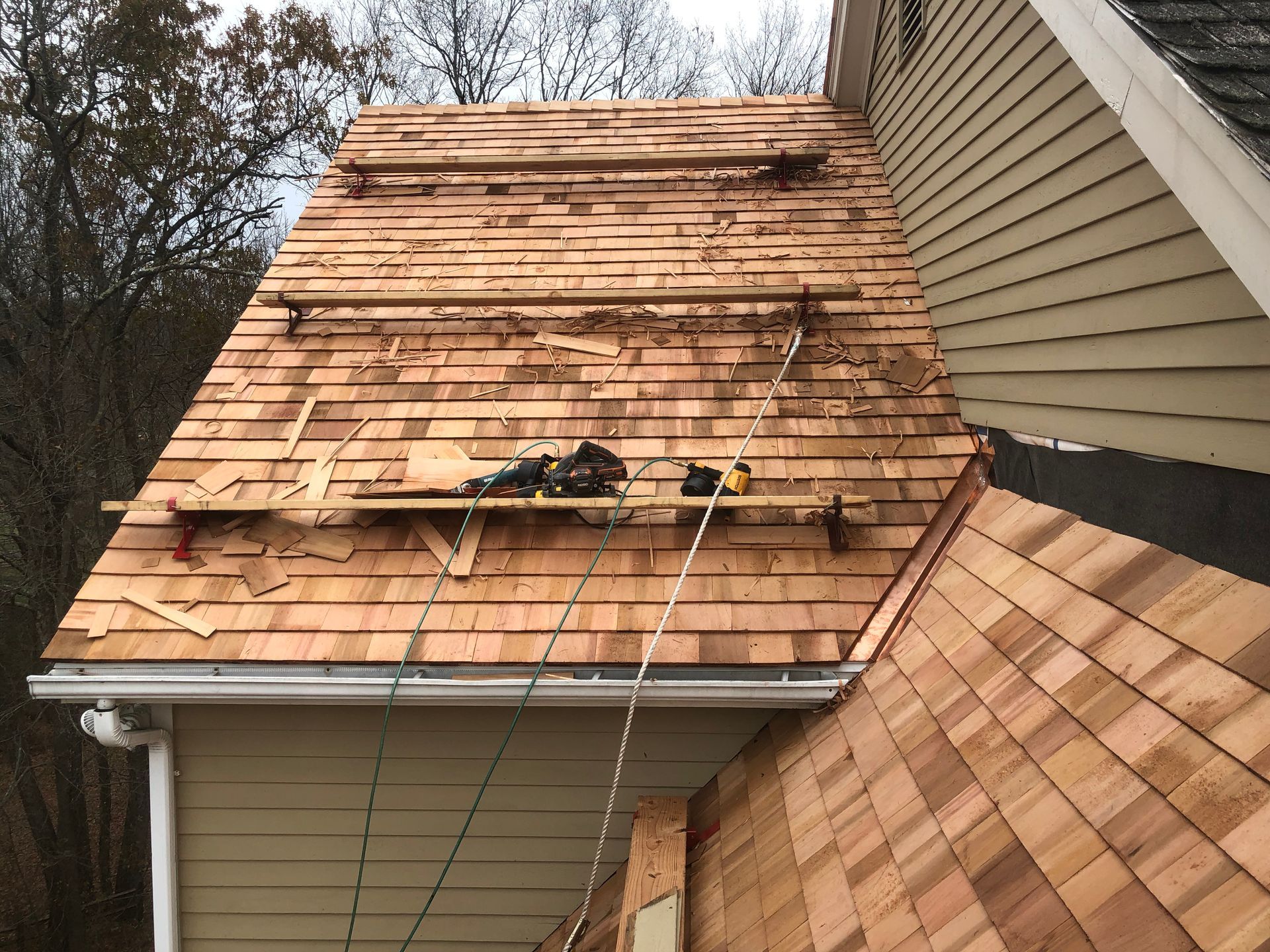 Roof partially covered with cedar shingles during construction, with tools and safety ropes visible.