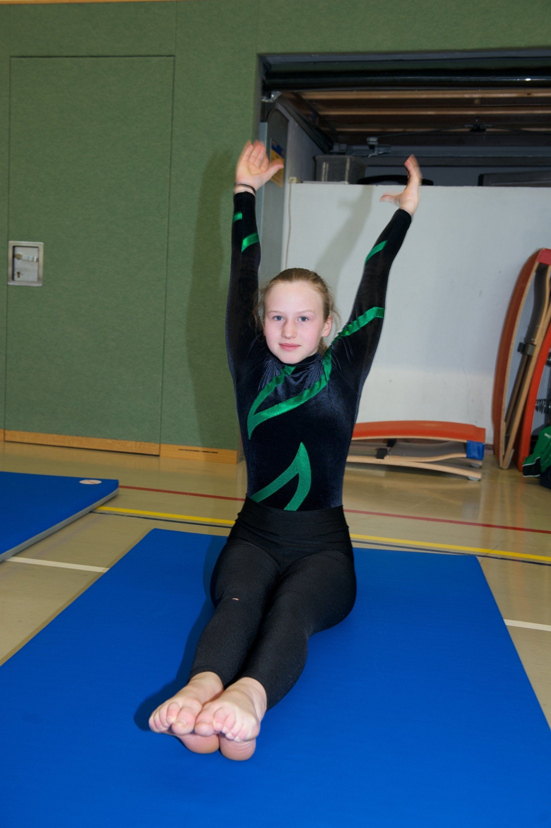 Ein junges Mädchen sitzt mit erhobenen Armen auf einer blauen Matte.