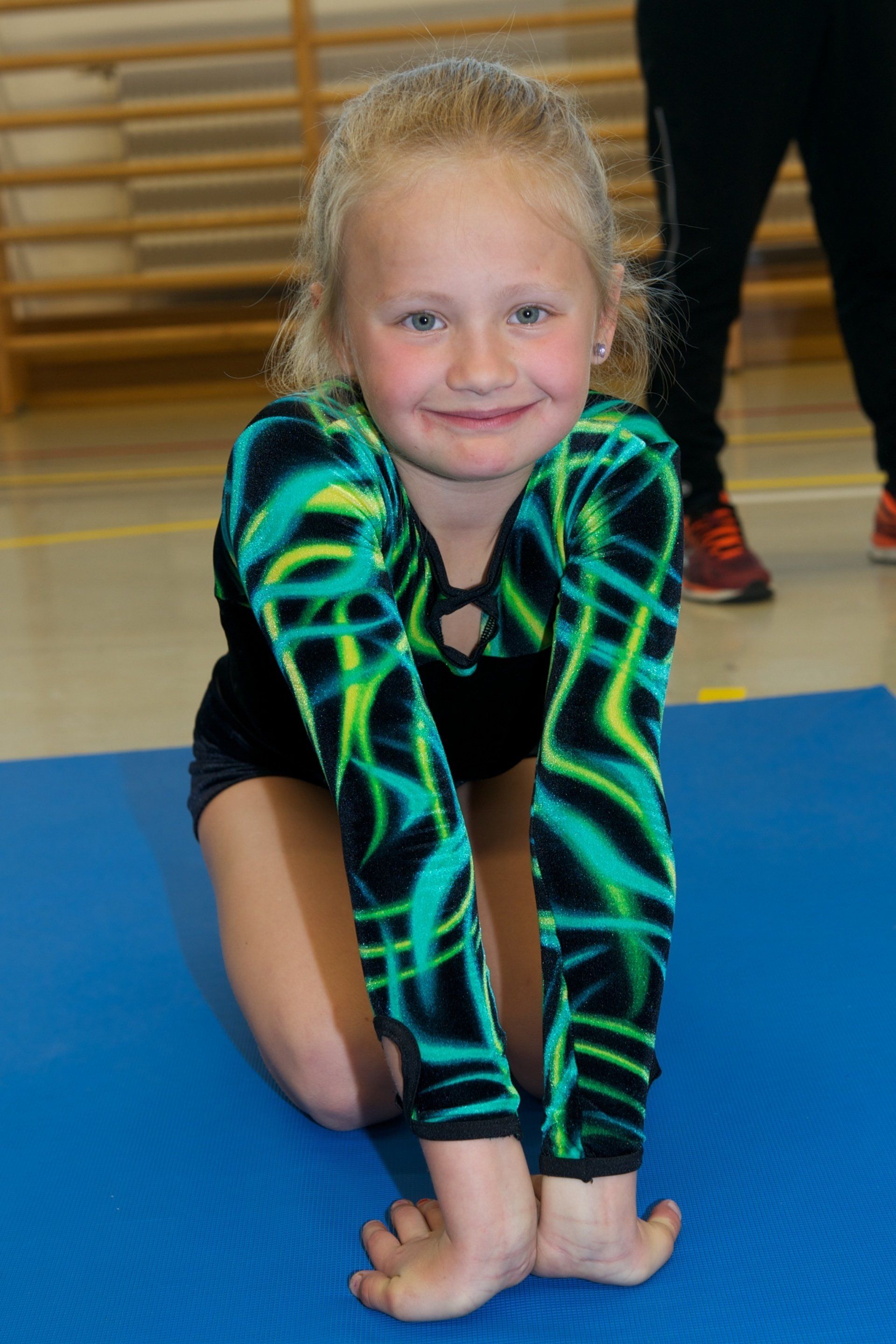 Ein junges Mädchen in einem grün-schwarzen Turnanzug sitzt auf einer blauen Matte.