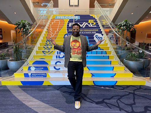 Dr. Adam Phyall stands in front of the staircase at IDEAcon. The stair stickers create an image on the bottom from left to right of the IDEA logo, the IDEAcon logo, a hand with the pointer finger pointing up the stairs, and on the top of the image are the IDEA lightbulb, the Rival 5 logo, and a text box that says, “Welcome.”