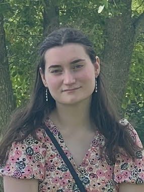 Woman with long brown hair, smiling, wearing floral top, and earrings; trees in background.