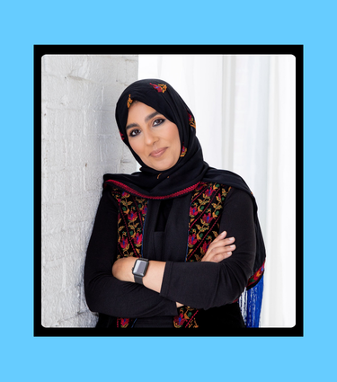 Woman in black headscarf and embroidered shawl, arms crossed, leaning against a white brick wall.