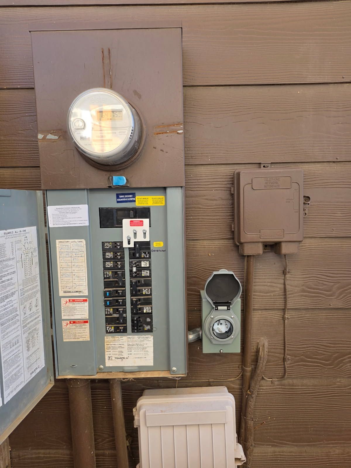 Electrical panel with meter, breakers, and conduits on a brown wooden wall.