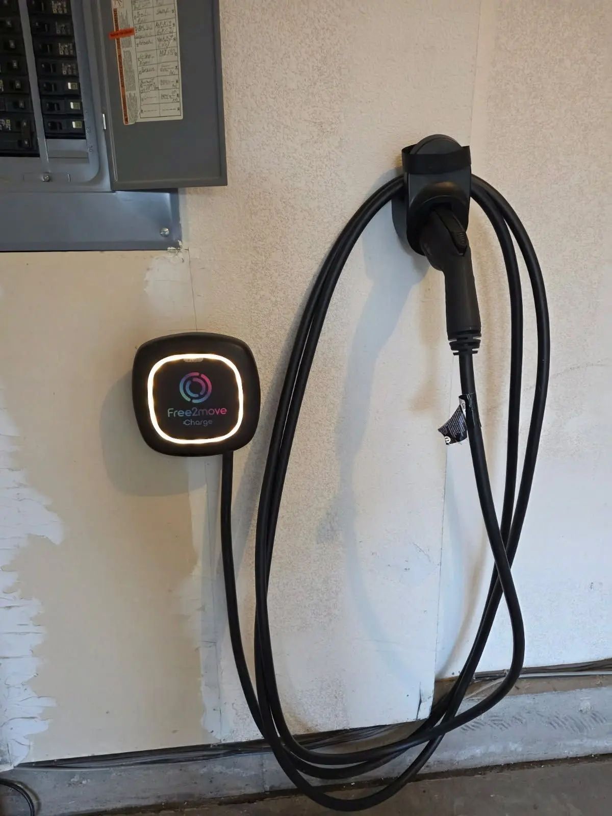 Electric vehicle charger mounted on a wall with charging cable. Electrical panel in the upper left.