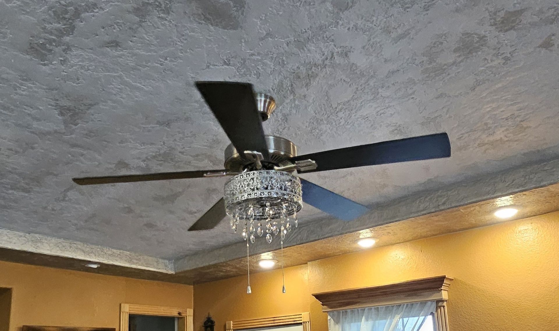 Ceiling fan with dark blades and crystal chandelier, in a room with textured ceiling.