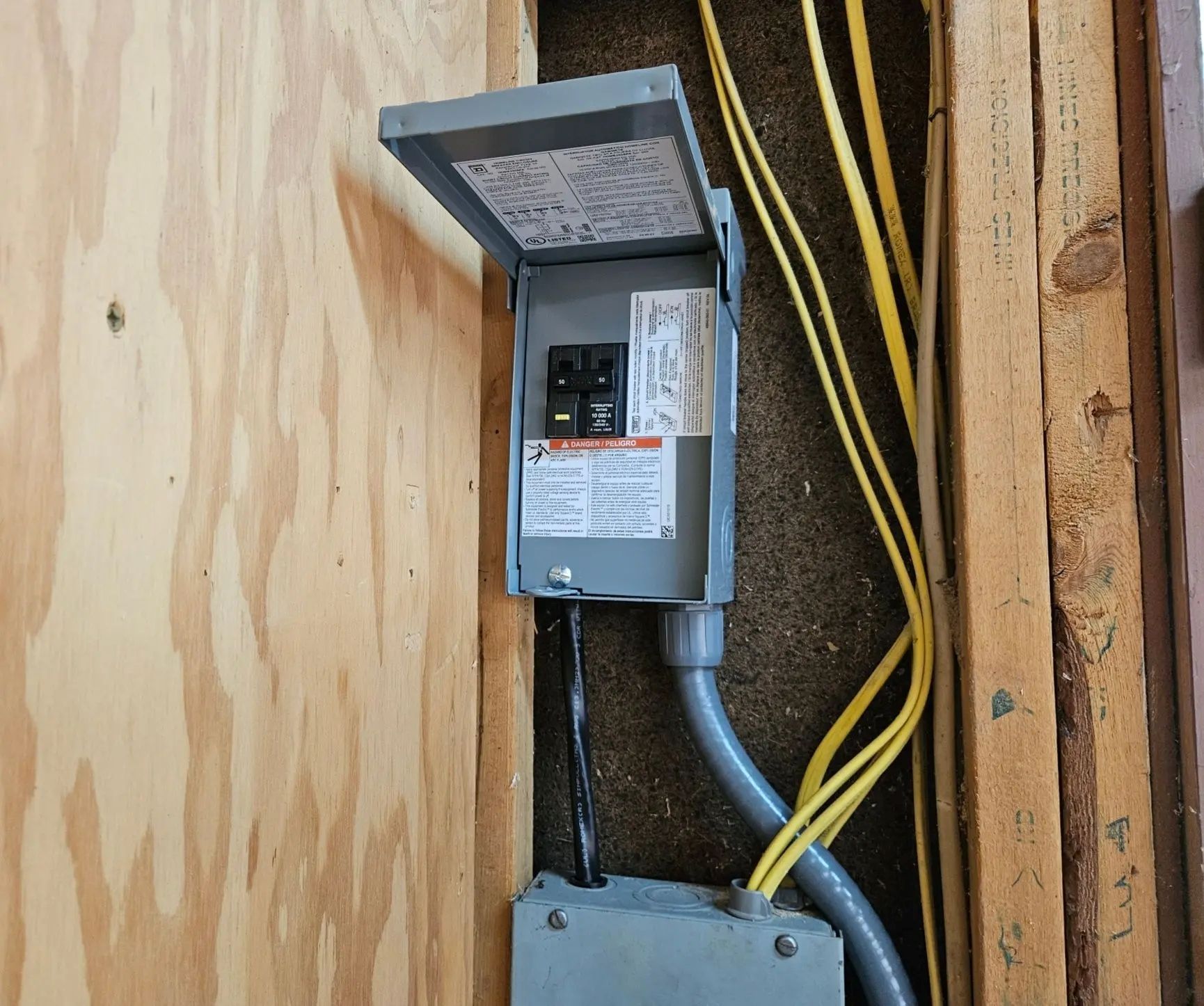 Electrical circuit breaker box mounted on wooden wall; gray box with door open, wires.