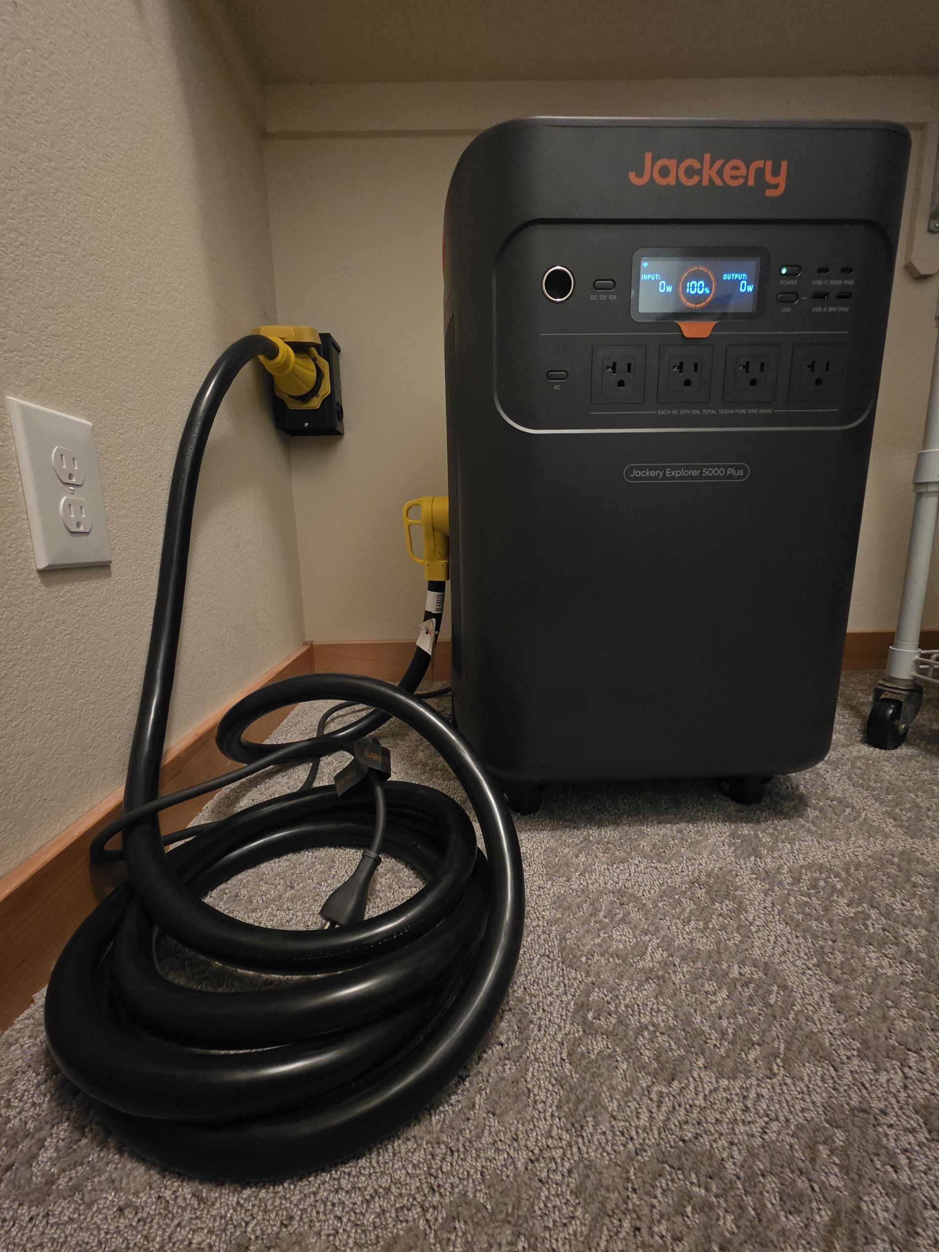 Black Jackery power station plugged into a wall outlet, with a coiled black cable on carpet.