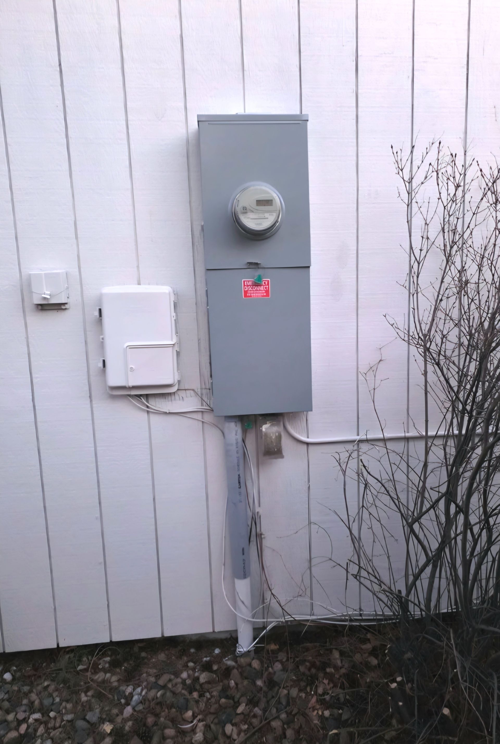 Gray electrical meter box and communication box on a white siding wall, with a shrub to the right.