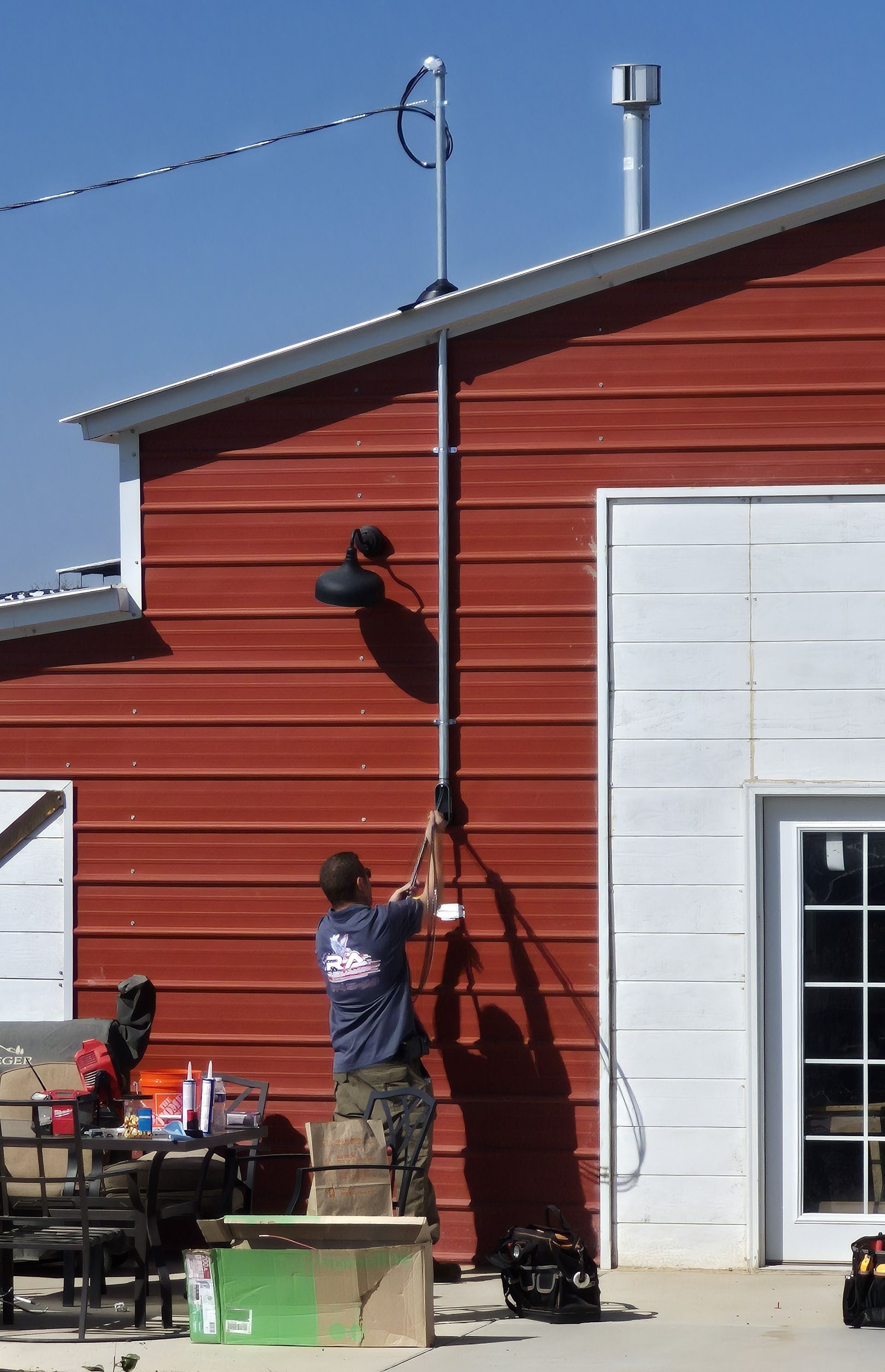 Man on a wooden crate installs something on a pole attached to a red barn. Outdoors, sunny.