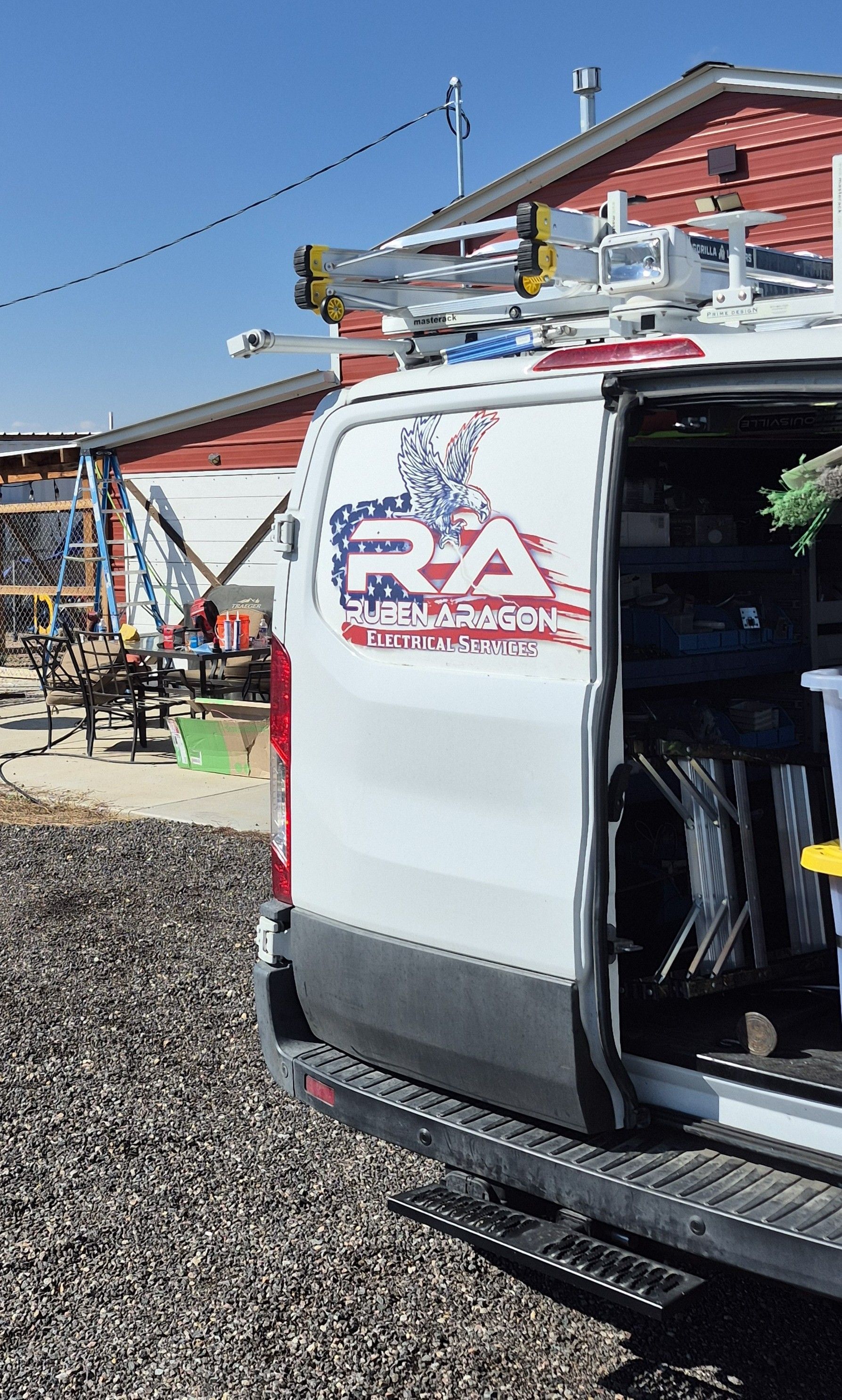 White work van with logo, parked near a red building on a sunny day. The rear door is open, showing the interior.