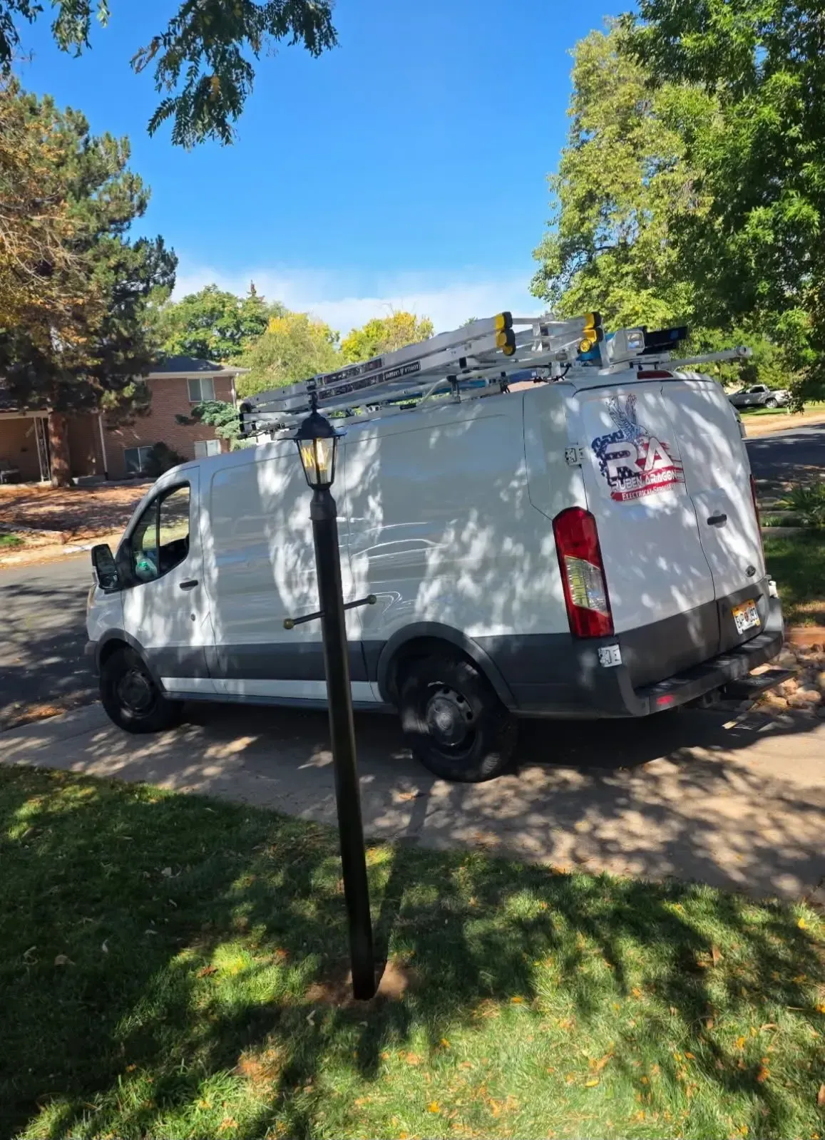 White work van with ladder on top parked on a residential street; blue sky, trees in the background.