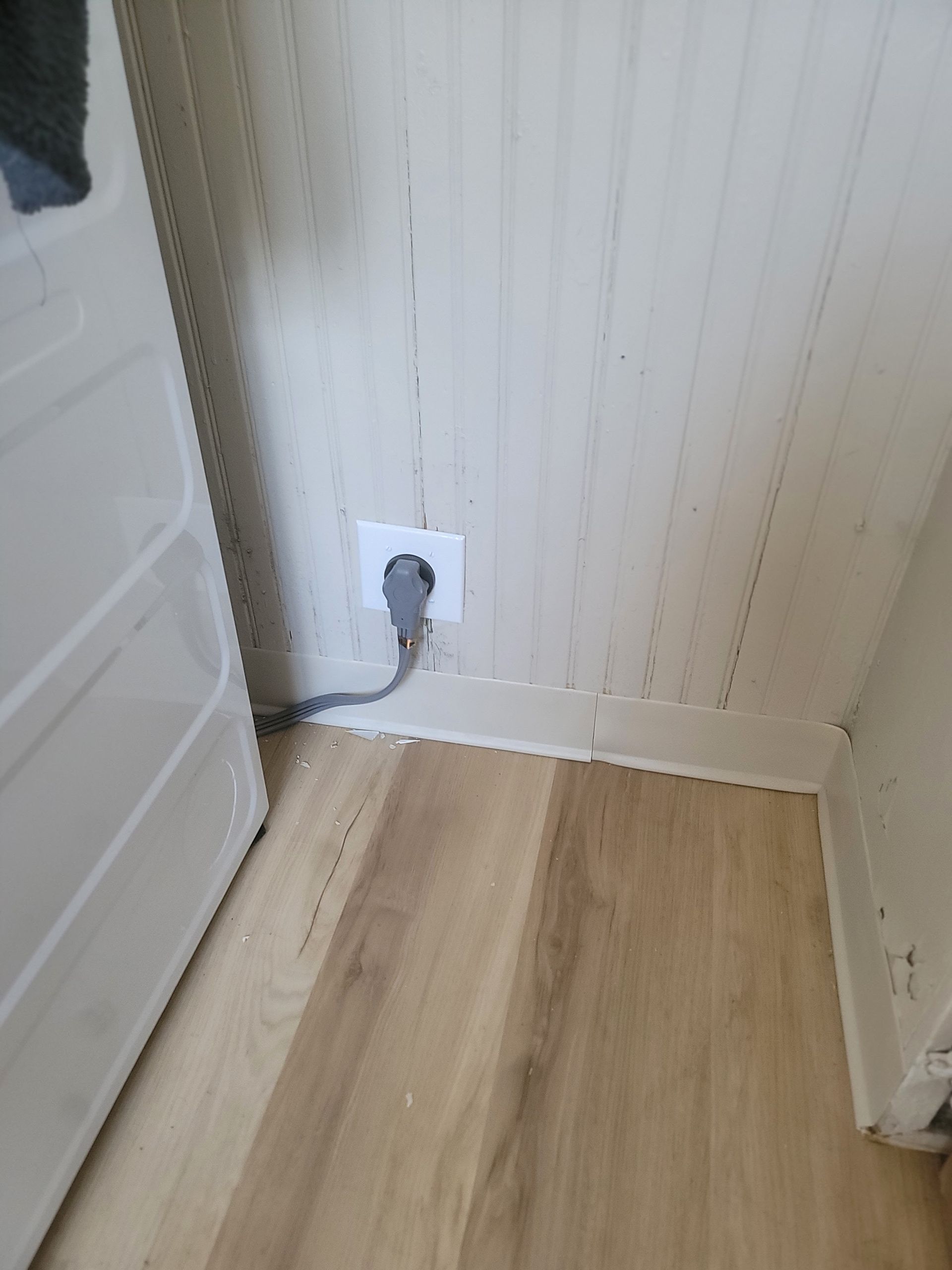 Electrical outlet on a wood-paneled wall, connected to a gray cord, near a white appliance and wood floor.
