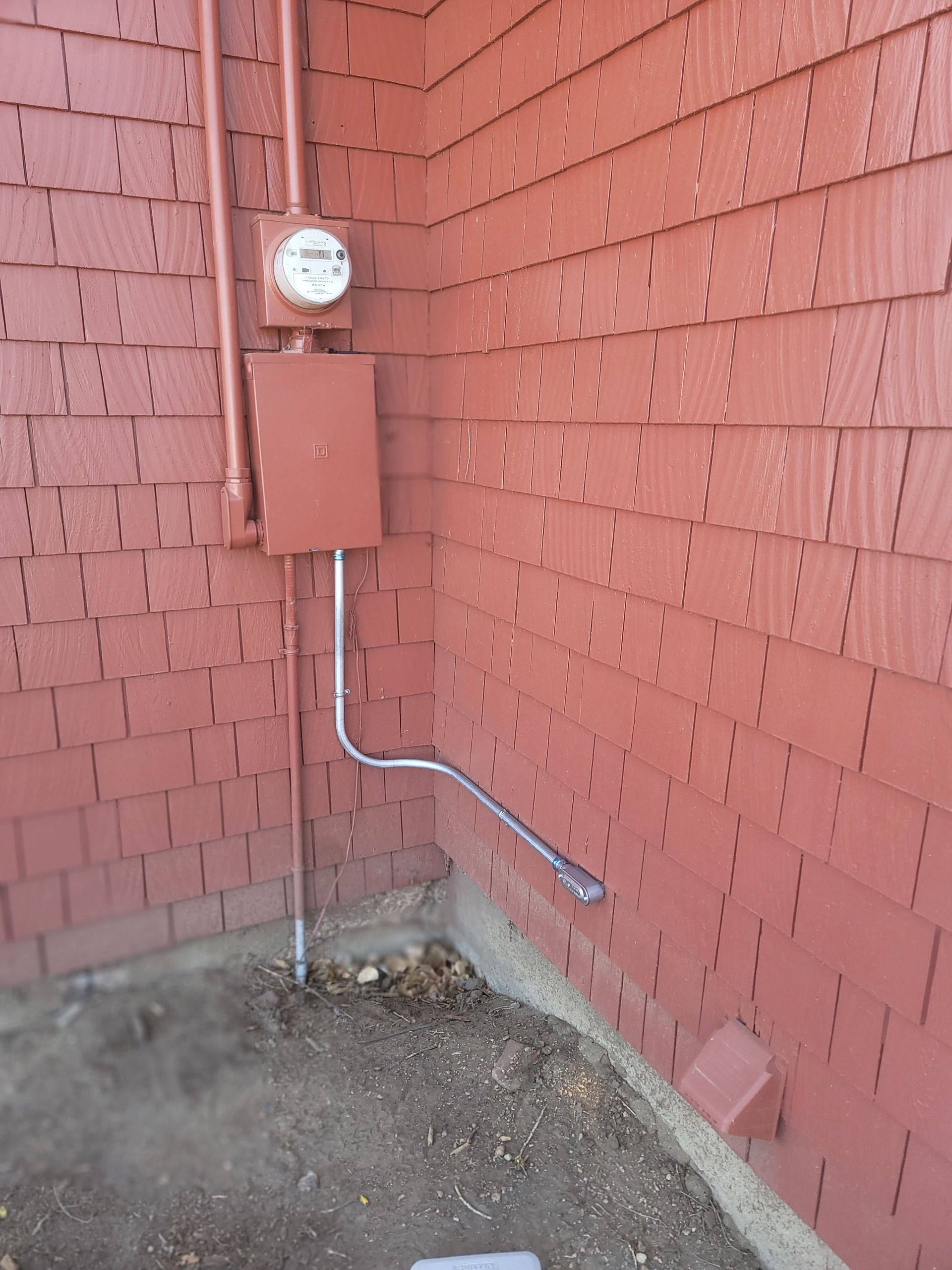 Brown electrical meter box and conduit on a red shingled building exterior.