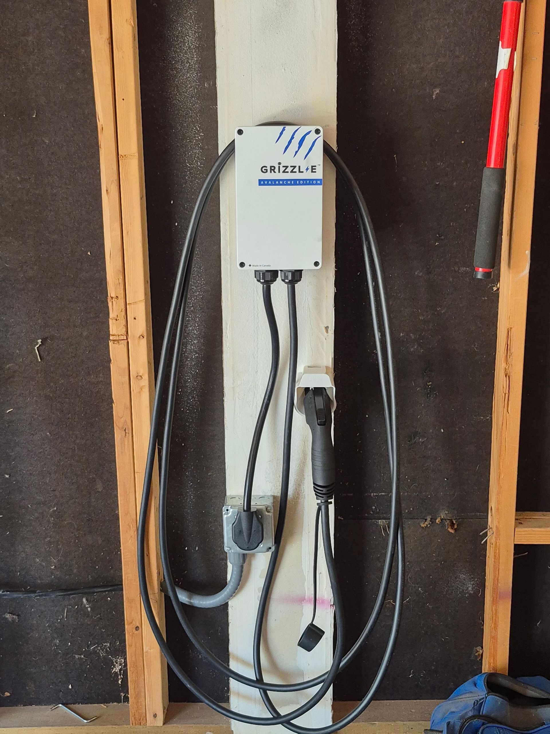 EV charger mounted on a wooden beam in a garage. Black cables connect to the charger, outlet, and vehicle port.