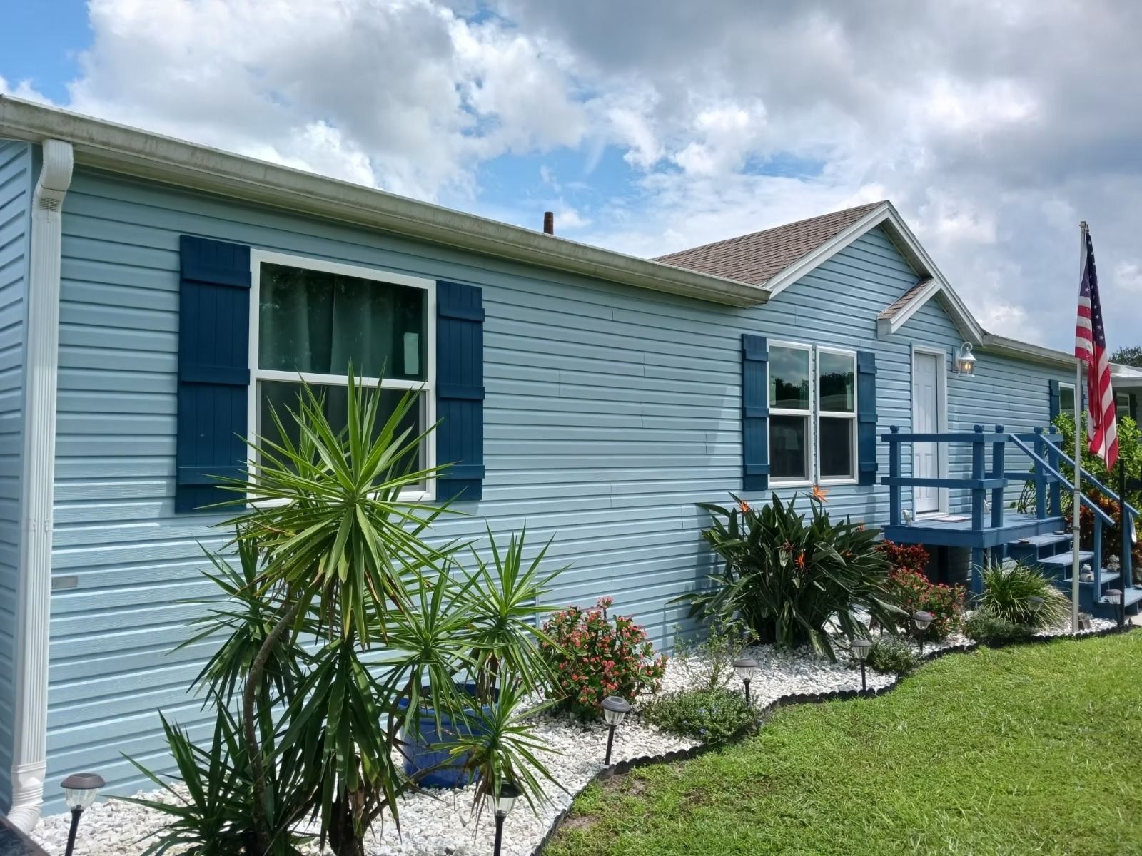 Light blue house with dark blue shutters, American flag, and landscaping under a cloudy sky.