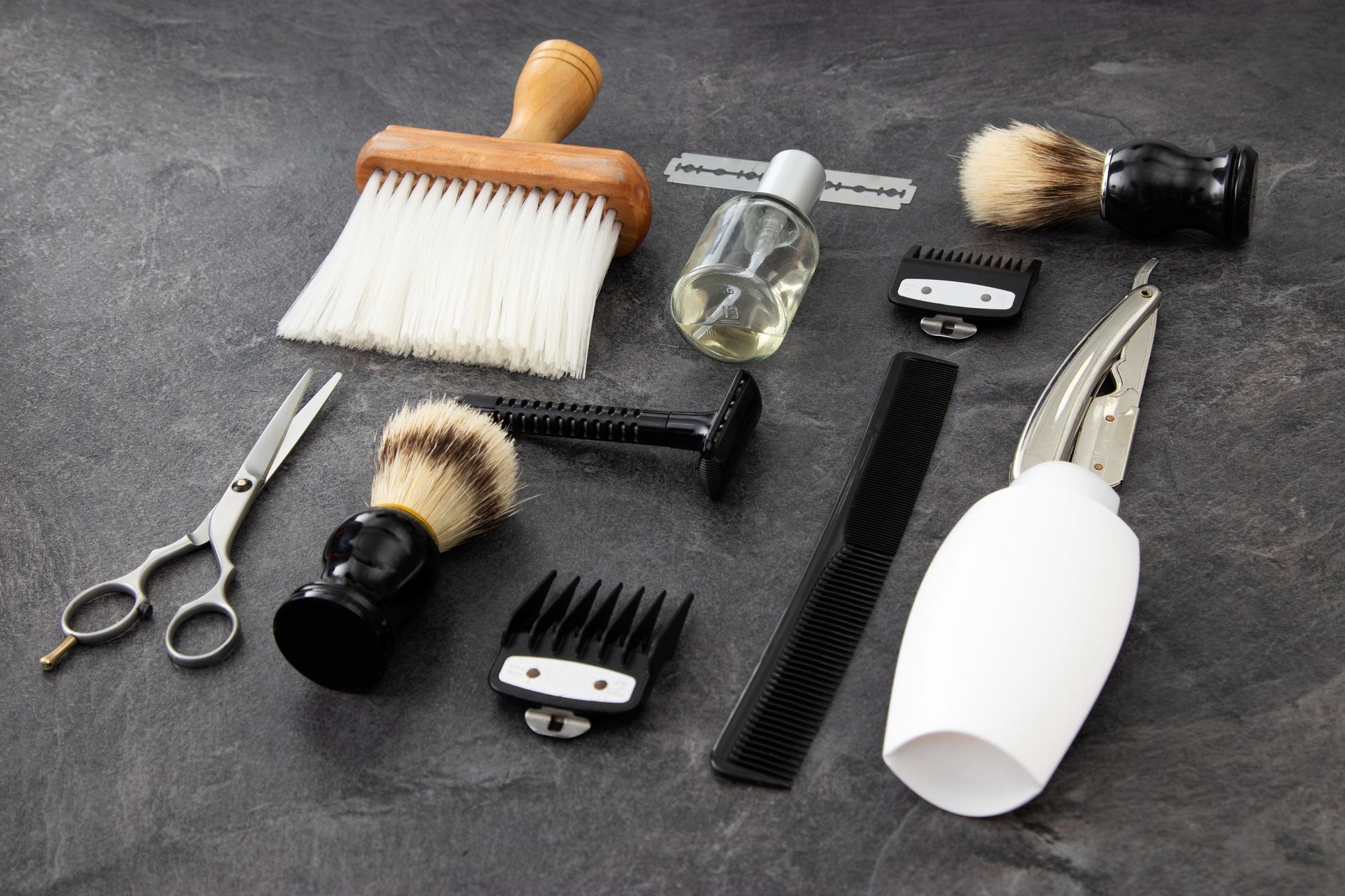 Herramientas de barbero dispuestas sobre una superficie oscura: tijeras, cepillos, cortapelos, maquinilla de afeitar, peine.