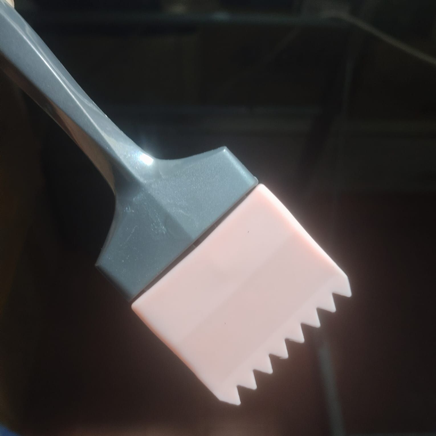 Una brocha de silicona con mango gris y punta rosa claro de borde dentado, sostenida sobre un fondo oscuro difuminado.