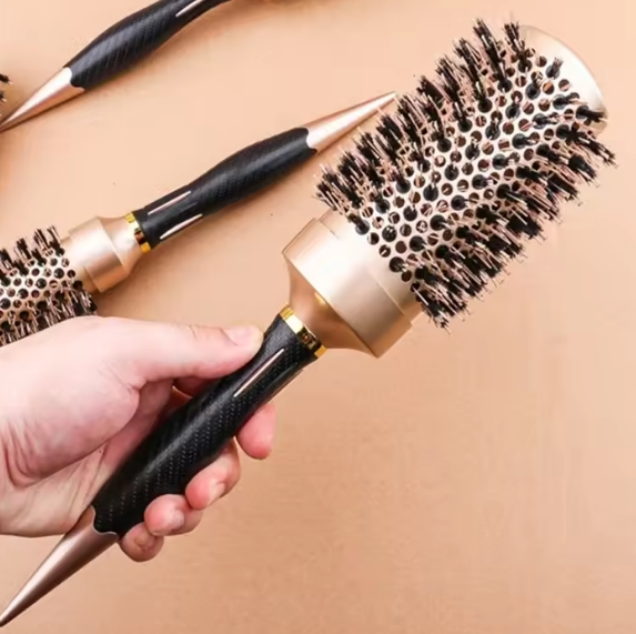 Una mano sostiene un cepillo redondo para el cabello con mango negro, base dorada y cerdas oscuras