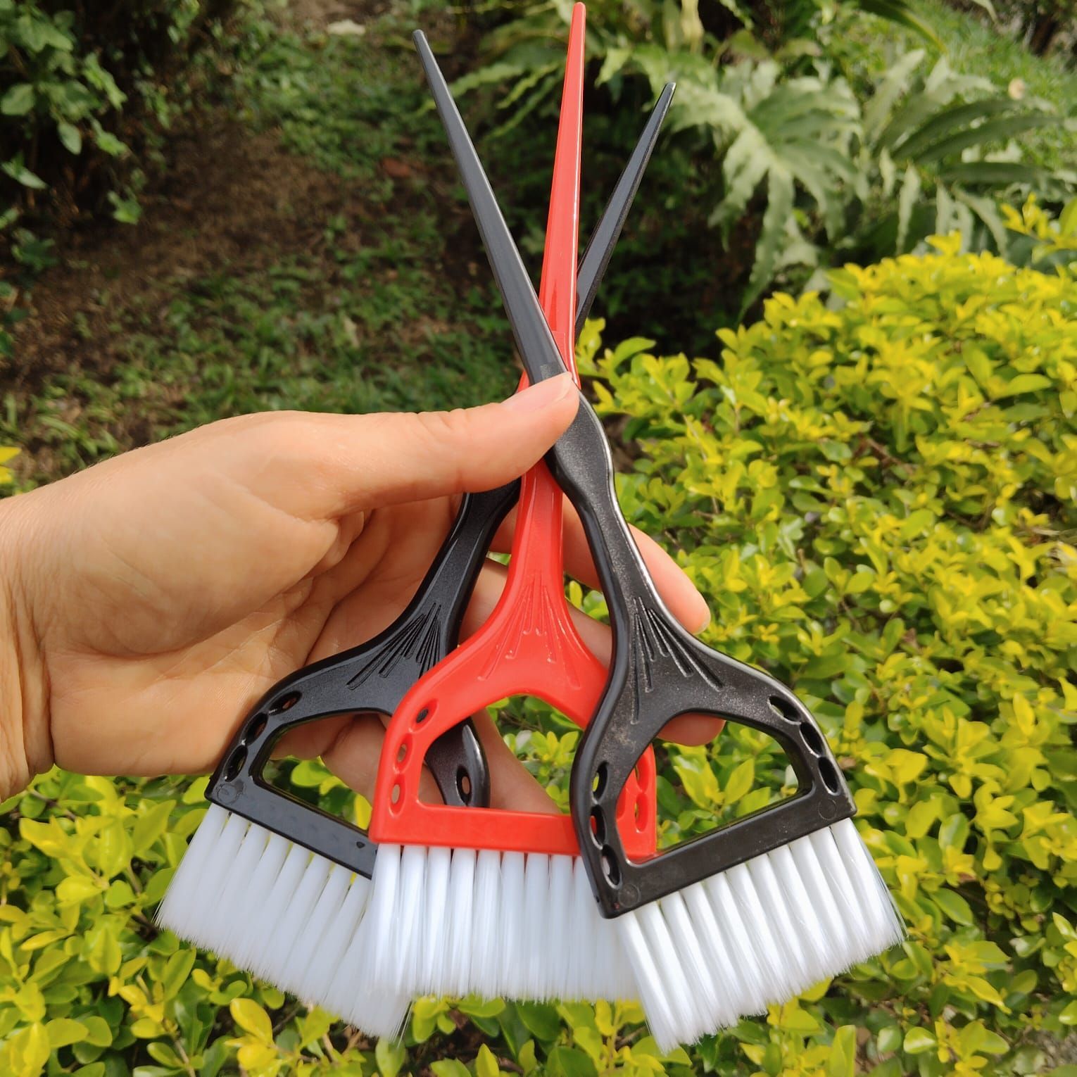 Una mano sostiene tres pinceles para teñir el cabello, con cerdas blancas, sobre un fondo de follaje verde.