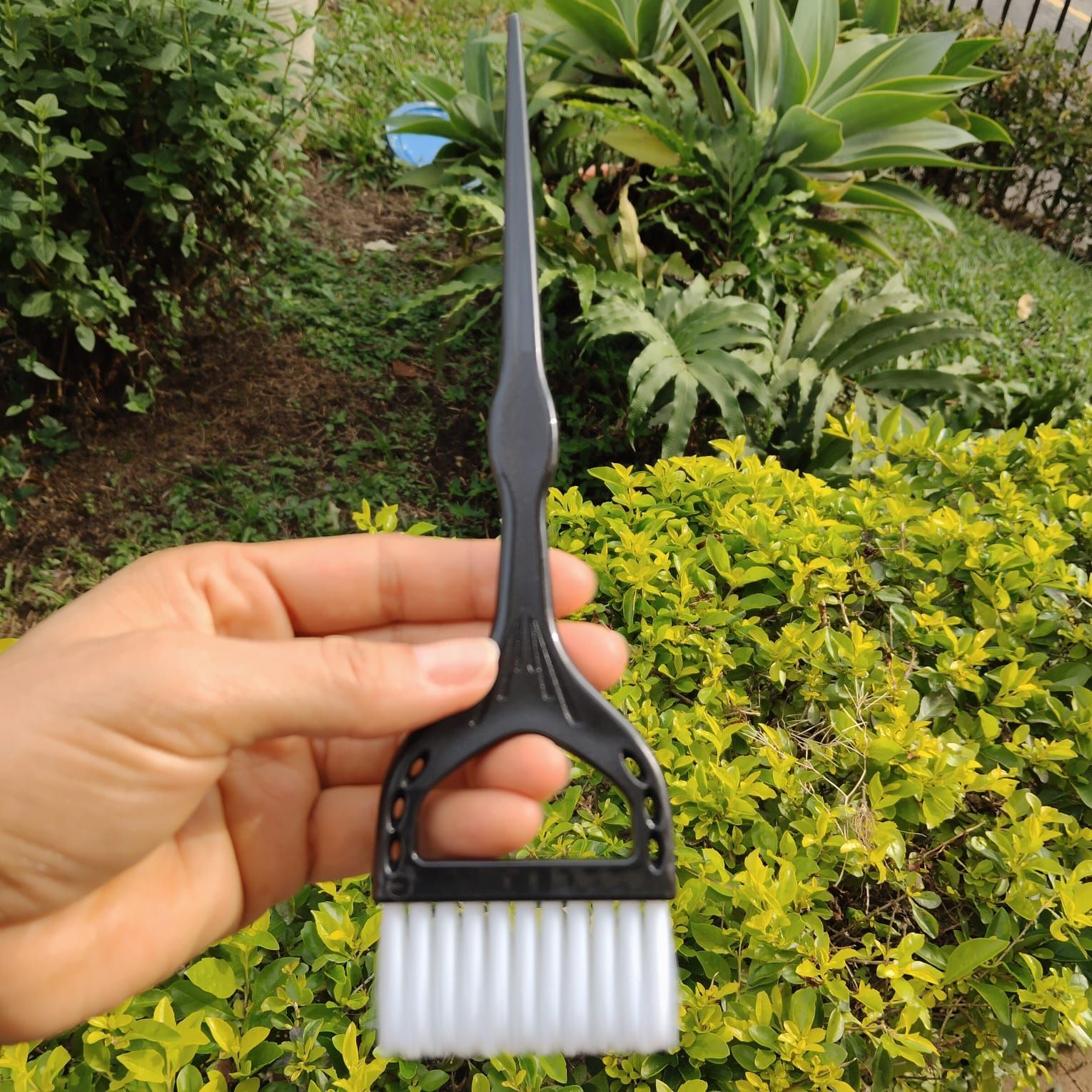 Una mano sostiene tres pinceles para teñir el cabello, con cerdas blancas, sobre un fondo de follaje verde.