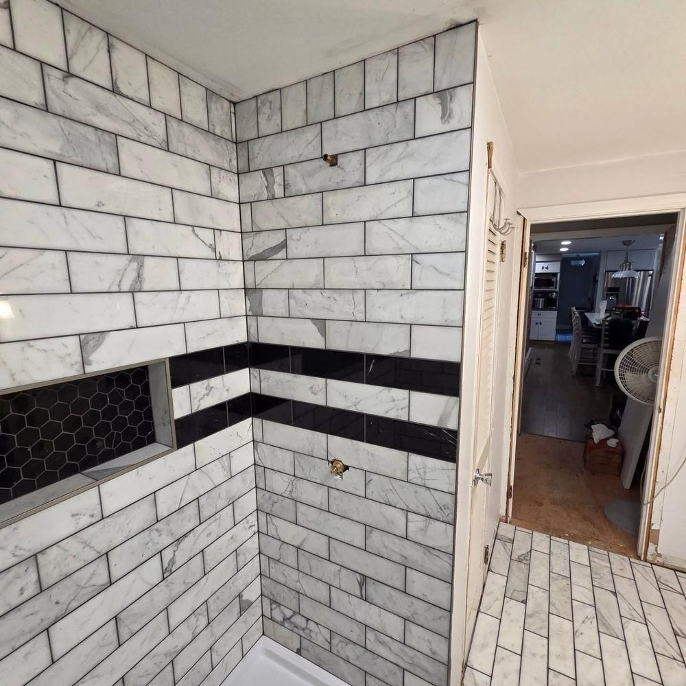 White subway-tile shower stall with black accent band and hex tile niche beside a tiled floor hallway