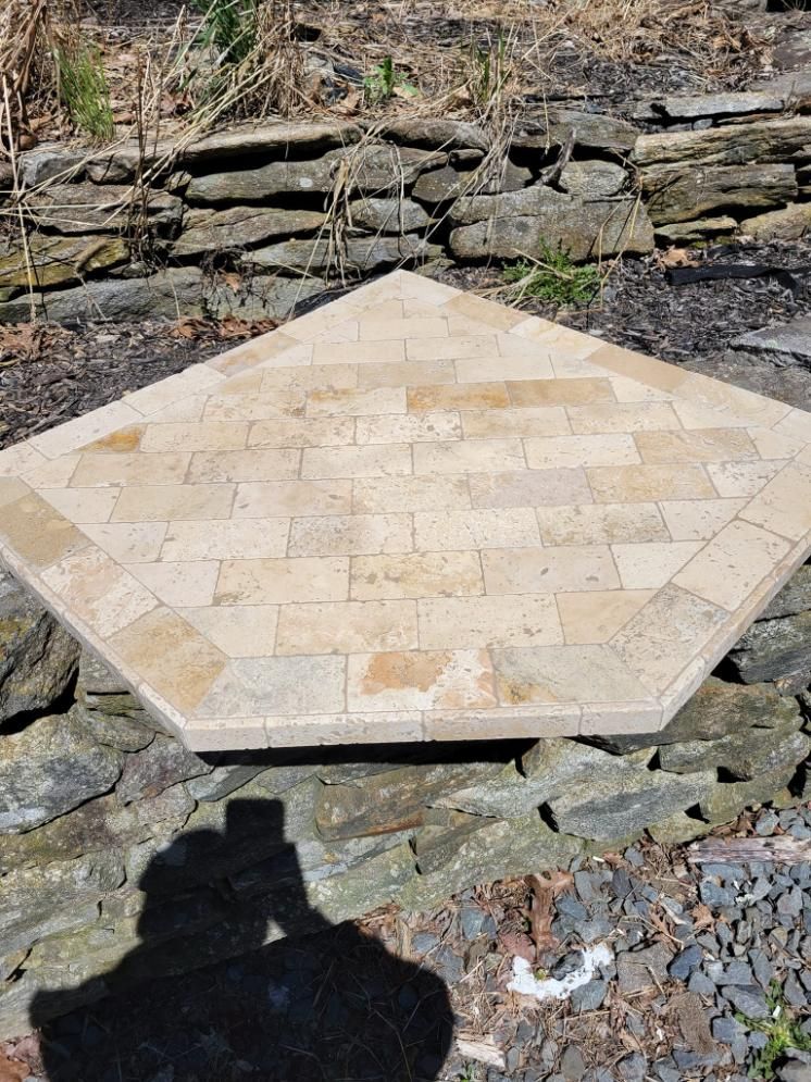 Beige hexagonal stone patio or table base on a rocky ground, with a stone wall behind.